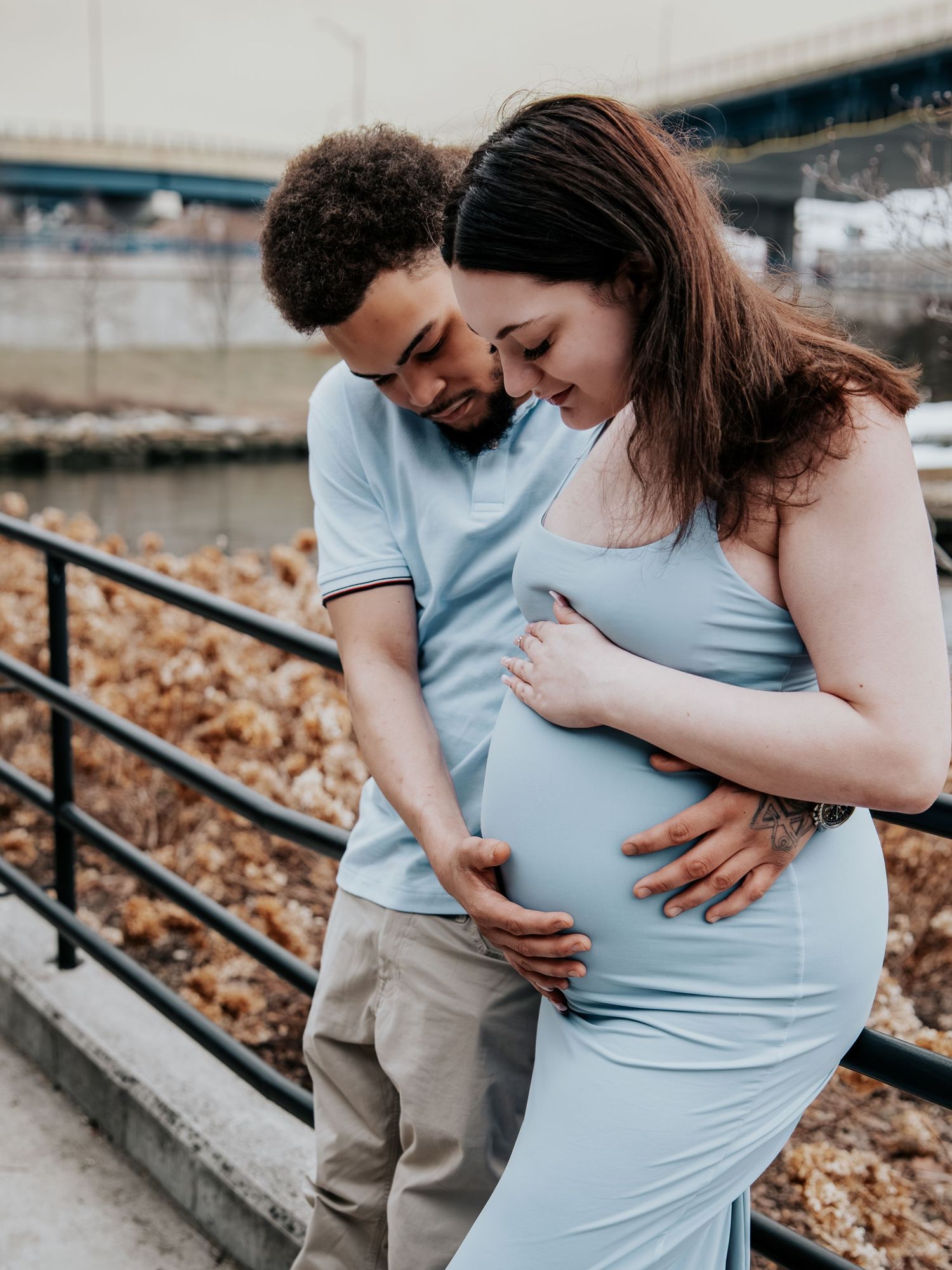 A couple tenderly cradles a pregnant belly outdoors near a railing, wearing matching light blue outfits during a maternity photoshoot.