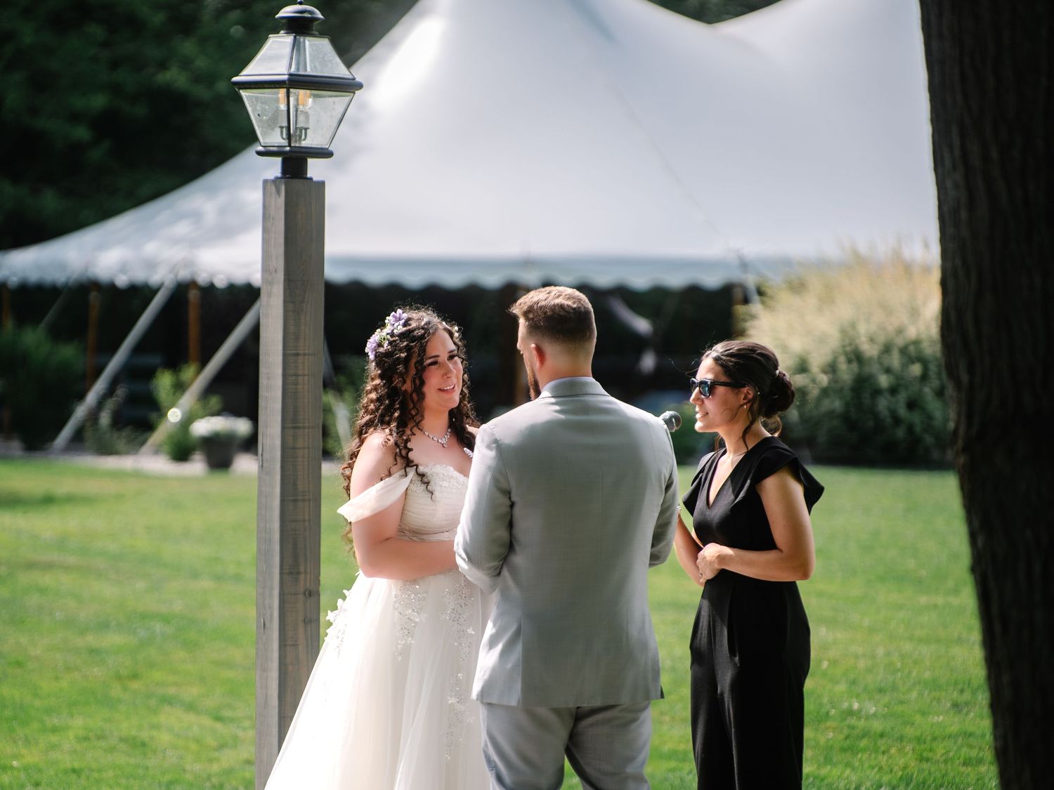 Outdoor wedding ceremony with bride in white gown, groom in gray suit, and officiant under a white tent.
