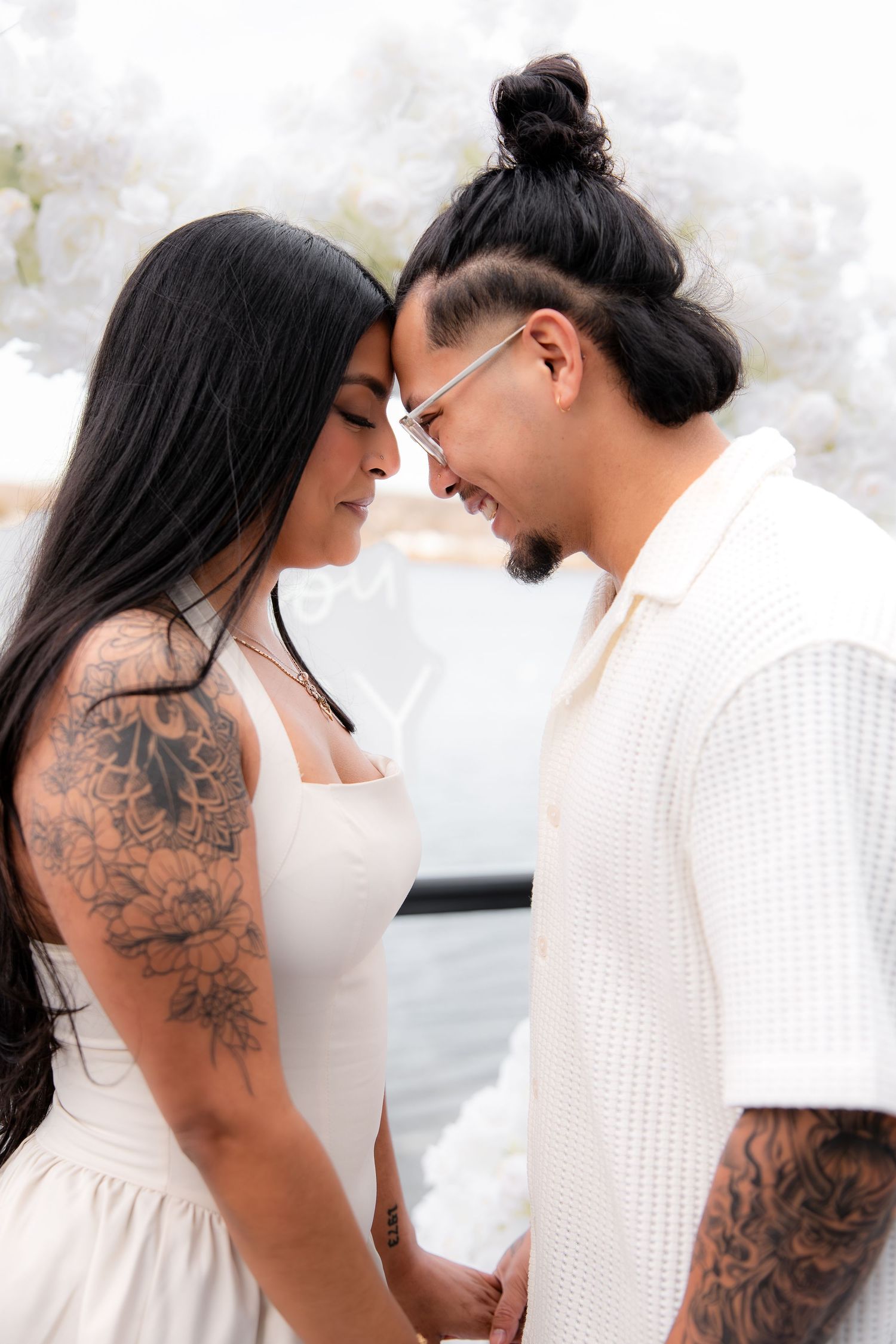 A tattooed couple in white outfits touch foreheads tenderly, holding hands in front of white floral backdrop.