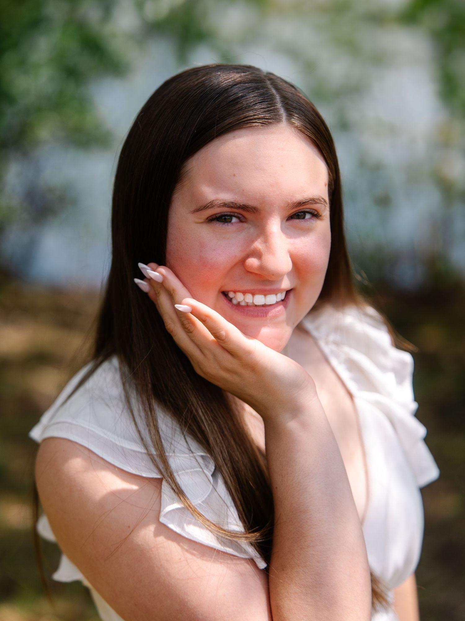 Smiling young woman with long dark hair in white top resting cheek on hand in sunny outdoor portrait.