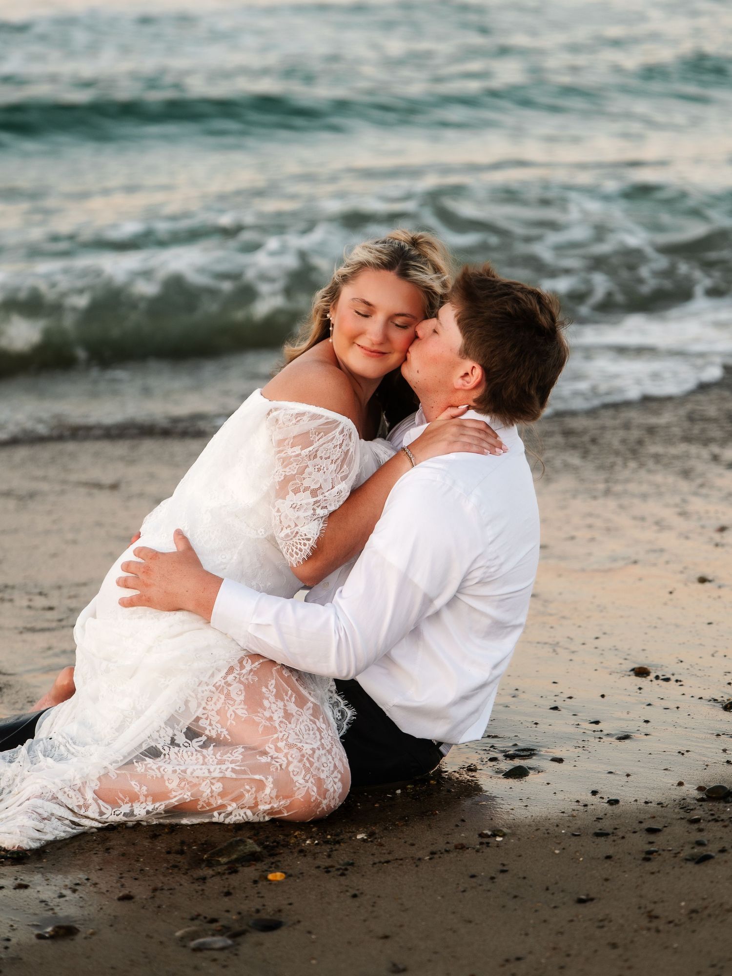 Smiling bride in white lace dress cuddled in groom's arms on sandy beach near ocean waves.