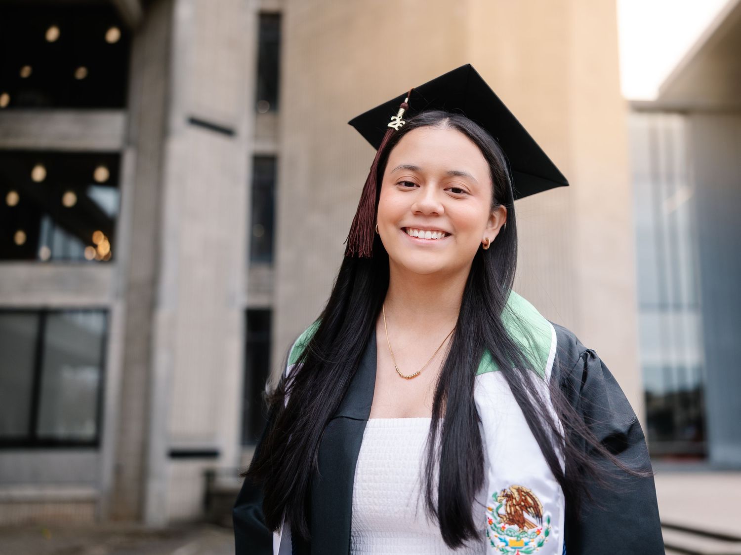 Smiling graduate in black cap and gown with Mexican flag stole poses outdoors.