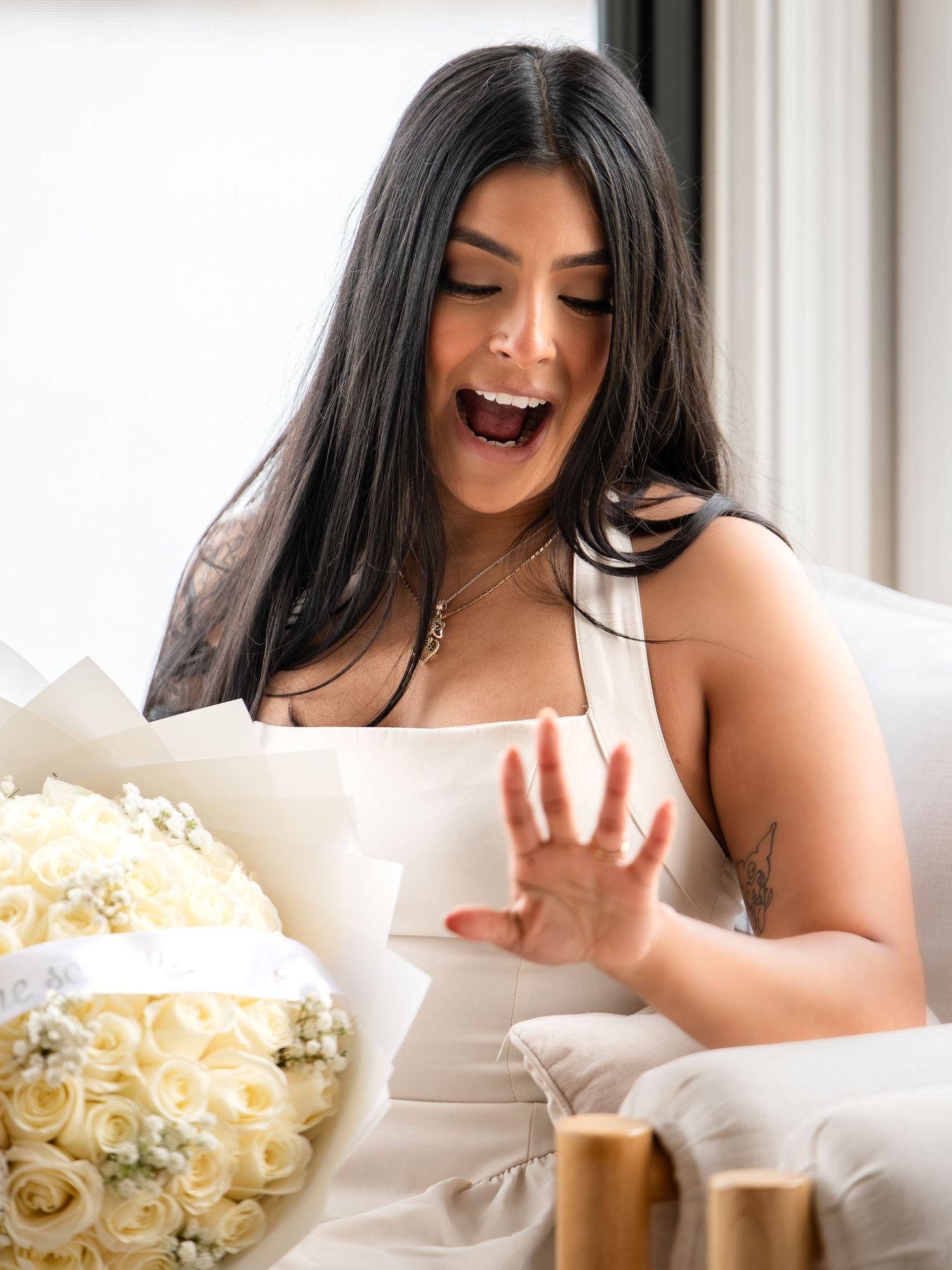 Excited woman looking at her engagement ring while holding white roses during a proposal moment.