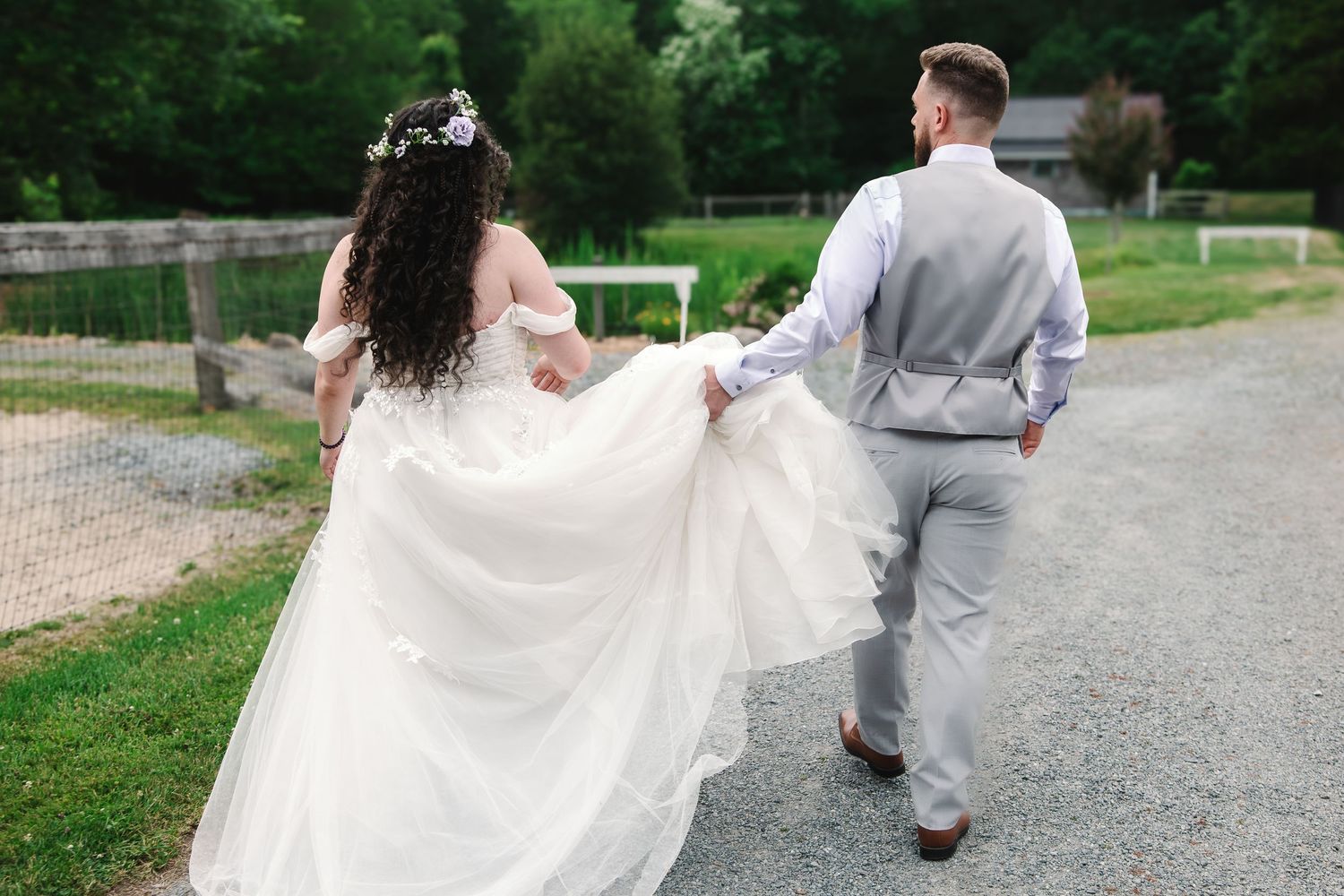 Bride in flowing white gown and groom in grey suit walking hand in hand along a farm pathway.