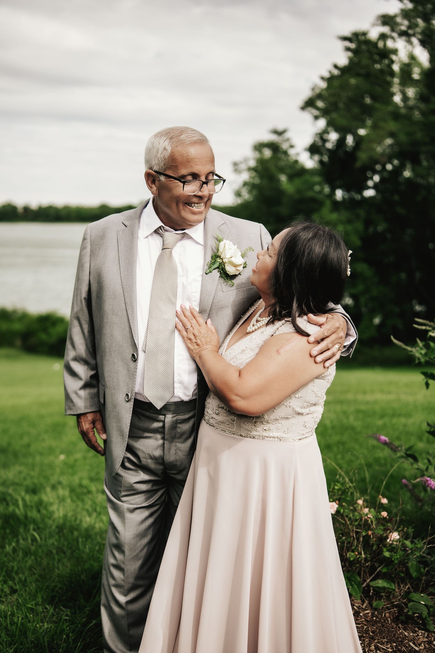 Older couple in wedding attire sharing a tender moment on a lush green lawn near a lake.