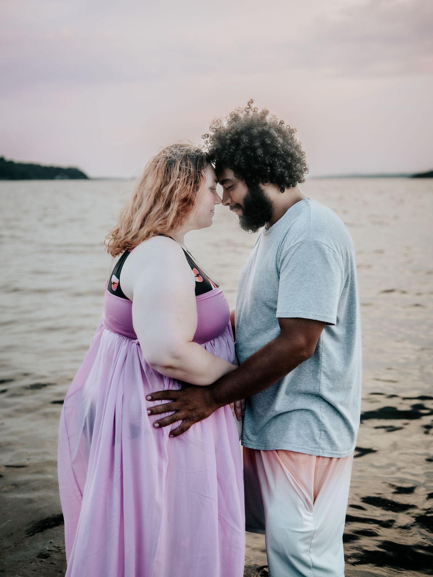 A couple shares a tender forehead touch while standing by a lake at sunset, the woman wearing a flowing lavender dress.