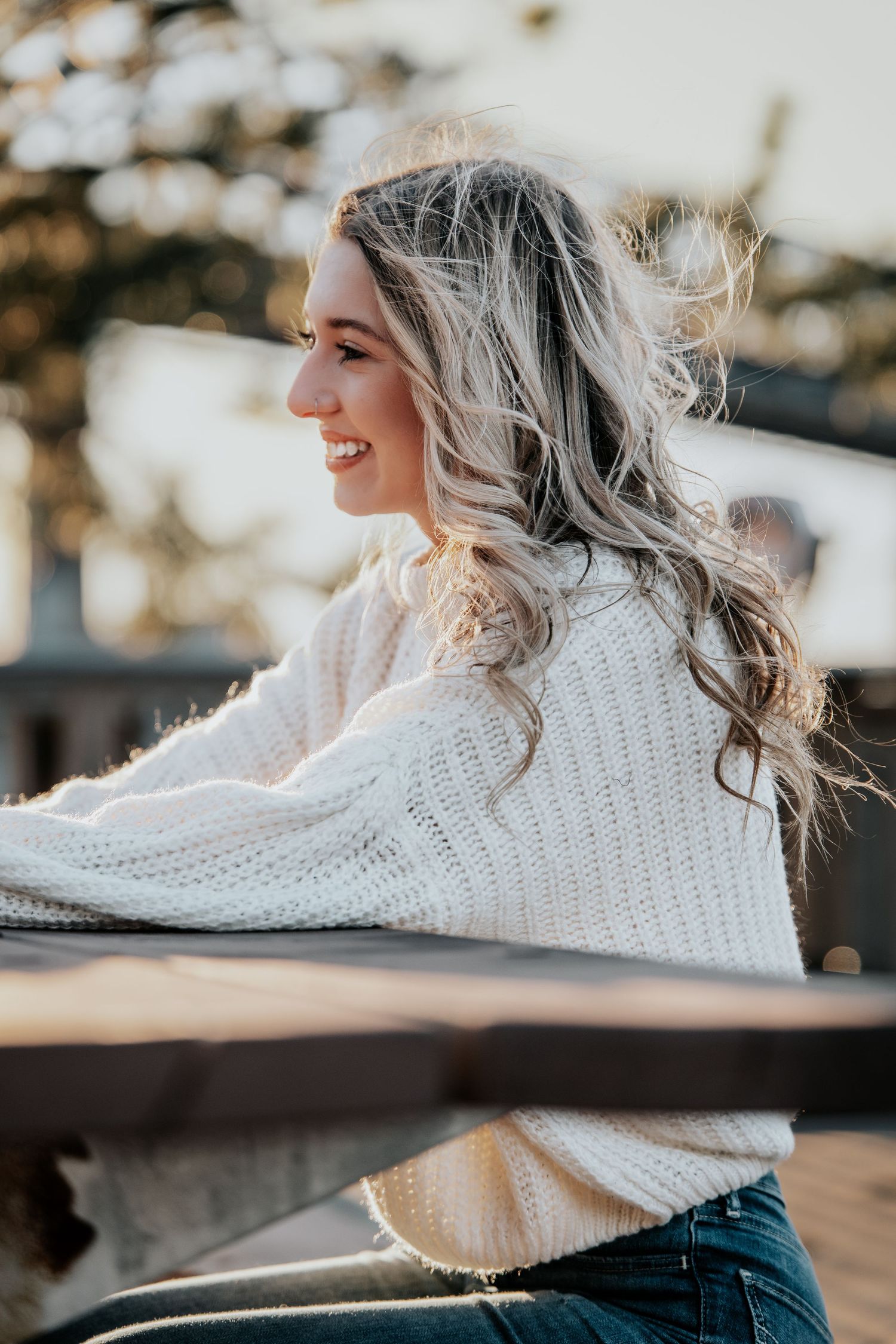 Smiling young woman with blonde hair wearing a white knit sweater sitting outdoors in warm sunlight.