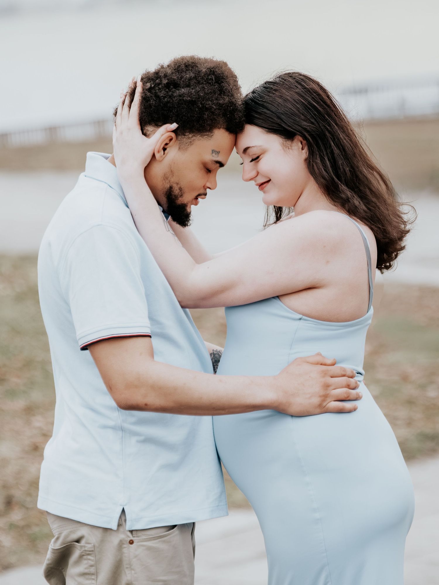 Couple sharing a tender forehead touch embrace outdoors, pregnant woman in blue dress with partner near waterfront.