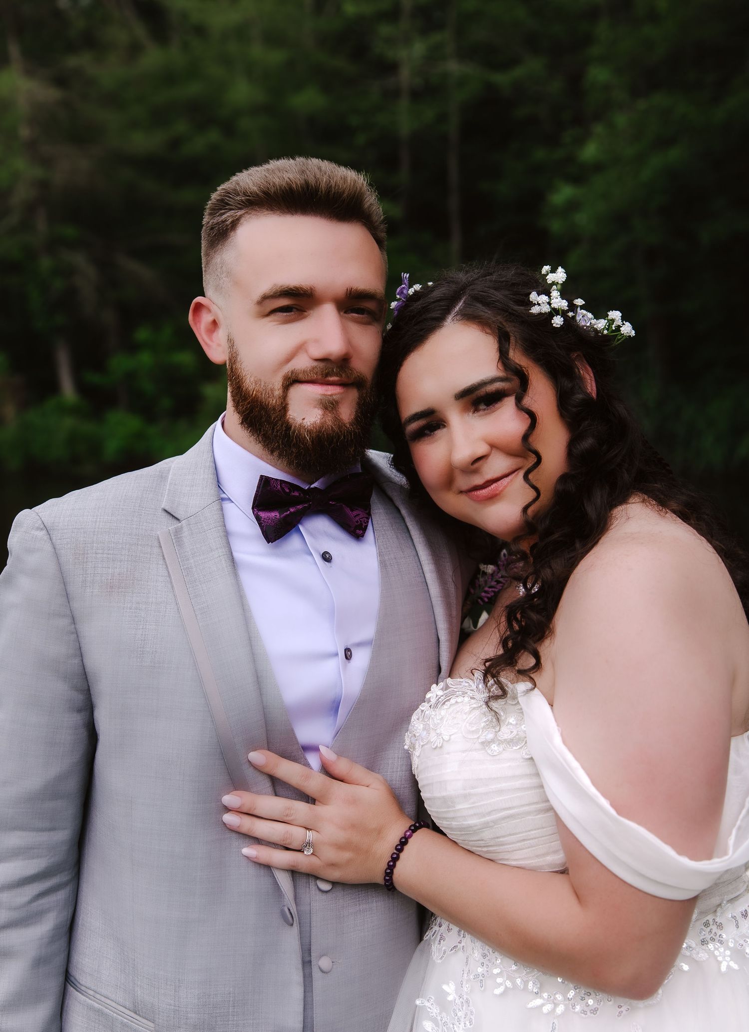 A couple in wedding attire poses outdoors, groom in gray suit with burgundy bow tie, bride in white off-shoulder gown with floral hair.