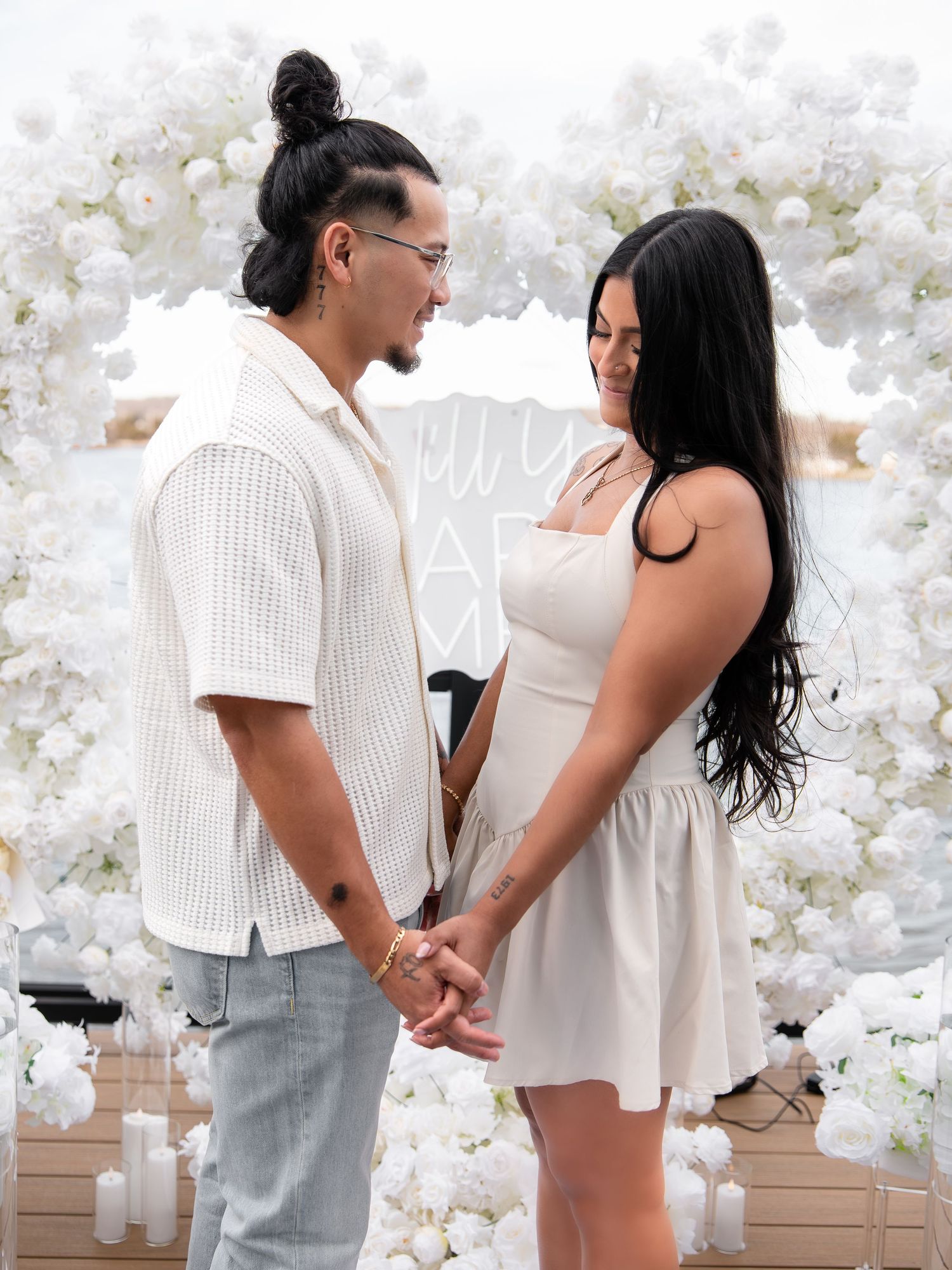 Couple holding hands smiling at each other in front of a white floral arch on a waterfront deck.