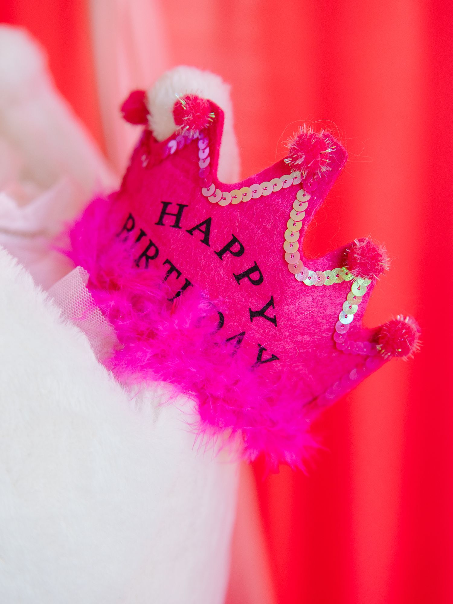 Hot pink sequined Happy Birthday crown with feather trim and pom poms held by a white fluffy stuffed animal.