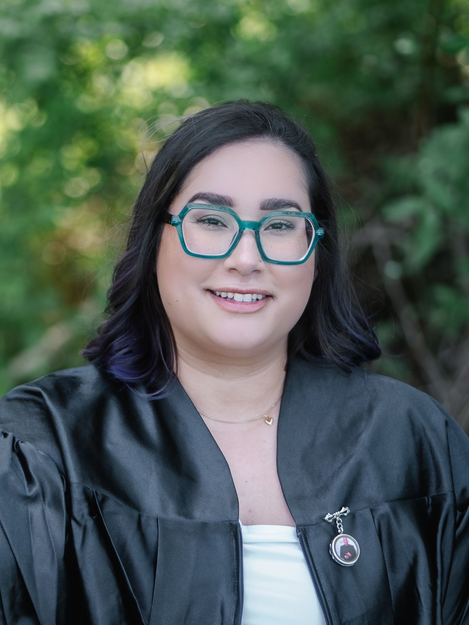 Smiling graduate in black gown and teal glasses posing for portrait with green foliage background.