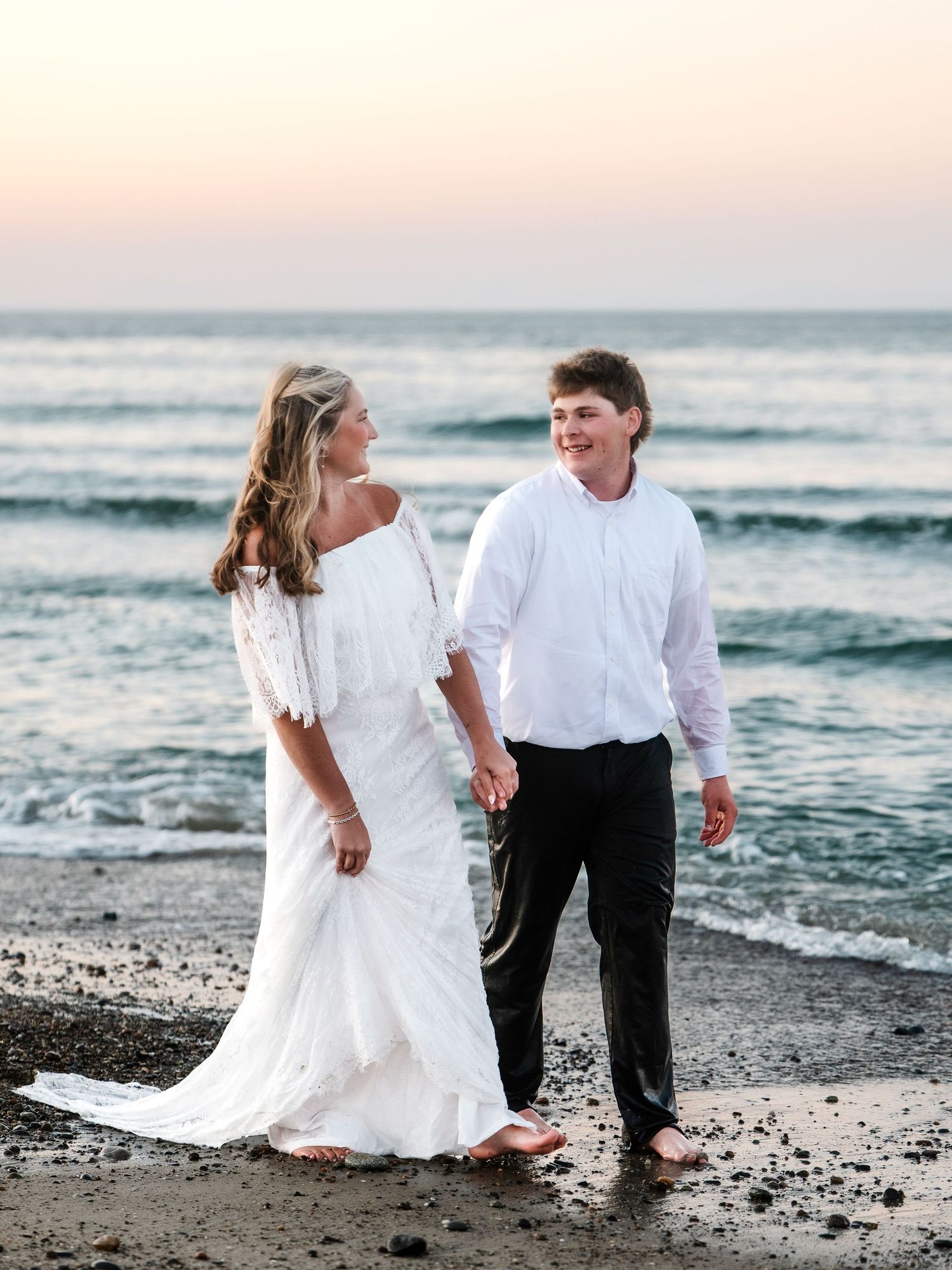 Bride and groom in white attire smiling at each other while walking on beach at sunset.