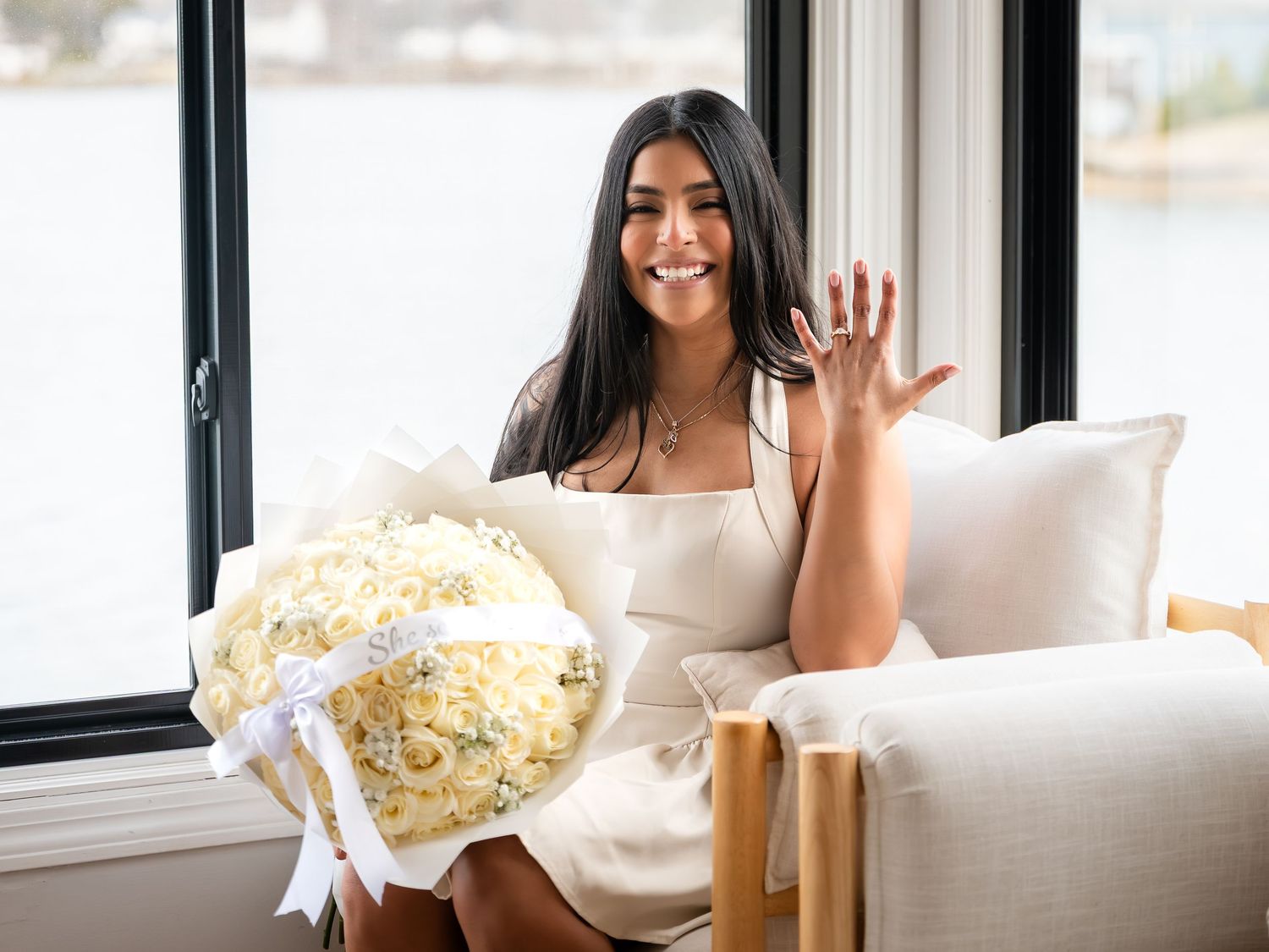 Smiling woman holding white roses bouquet showing engagement ring by waterfront window.
