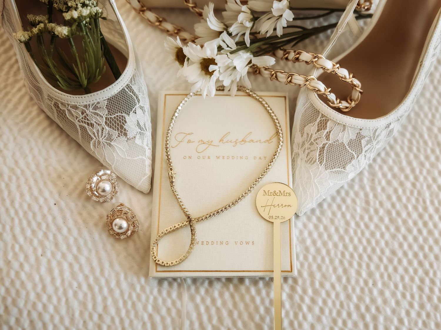 Elegant wedding flat lay with lace bridal heels, gold necklace, pearl earrings, cake topper, and a husband card on white fabric.