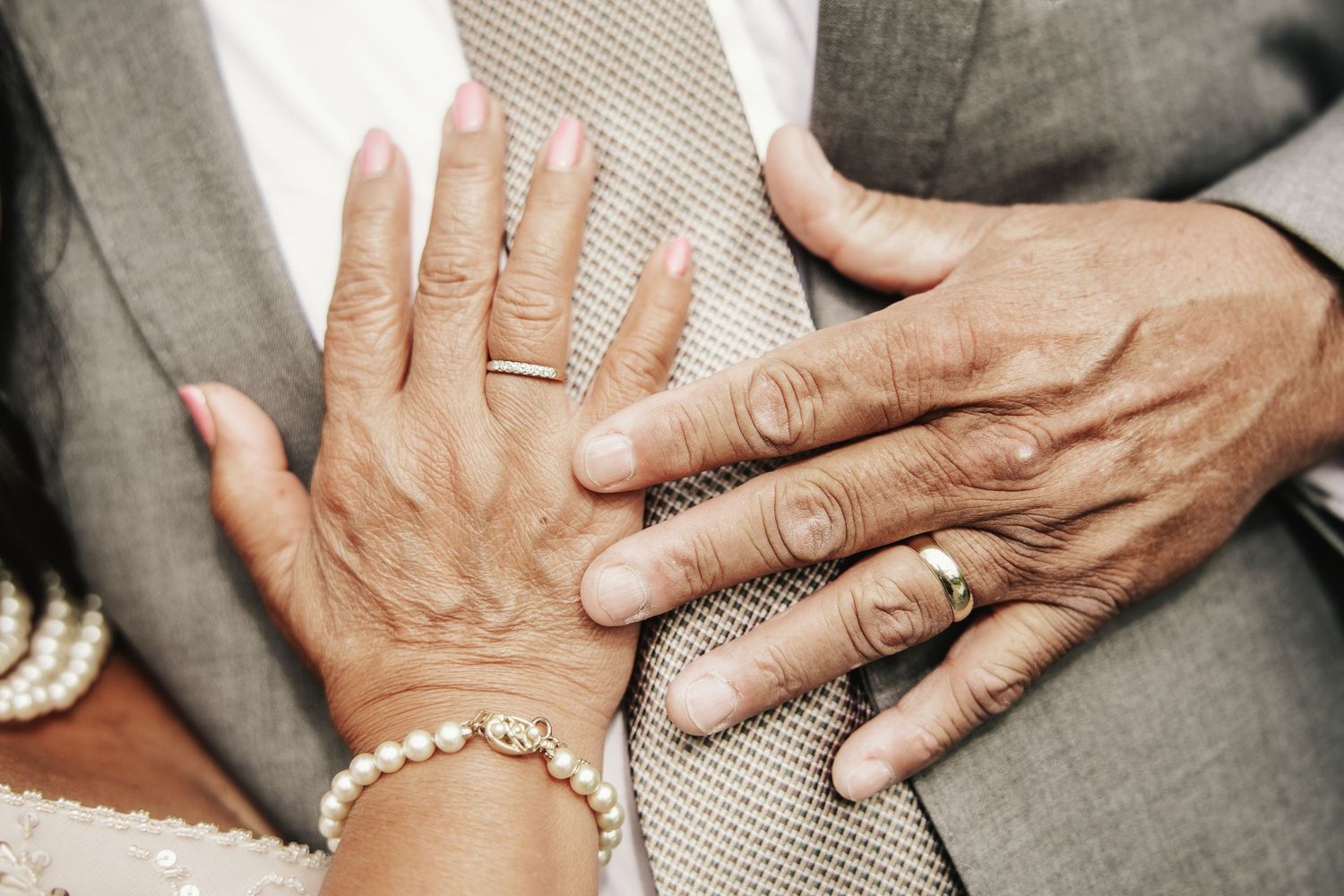 Elderly couple's hands showing wedding rings, pearl bracelet, and gray suit, symbolizing lasting love and marriage.