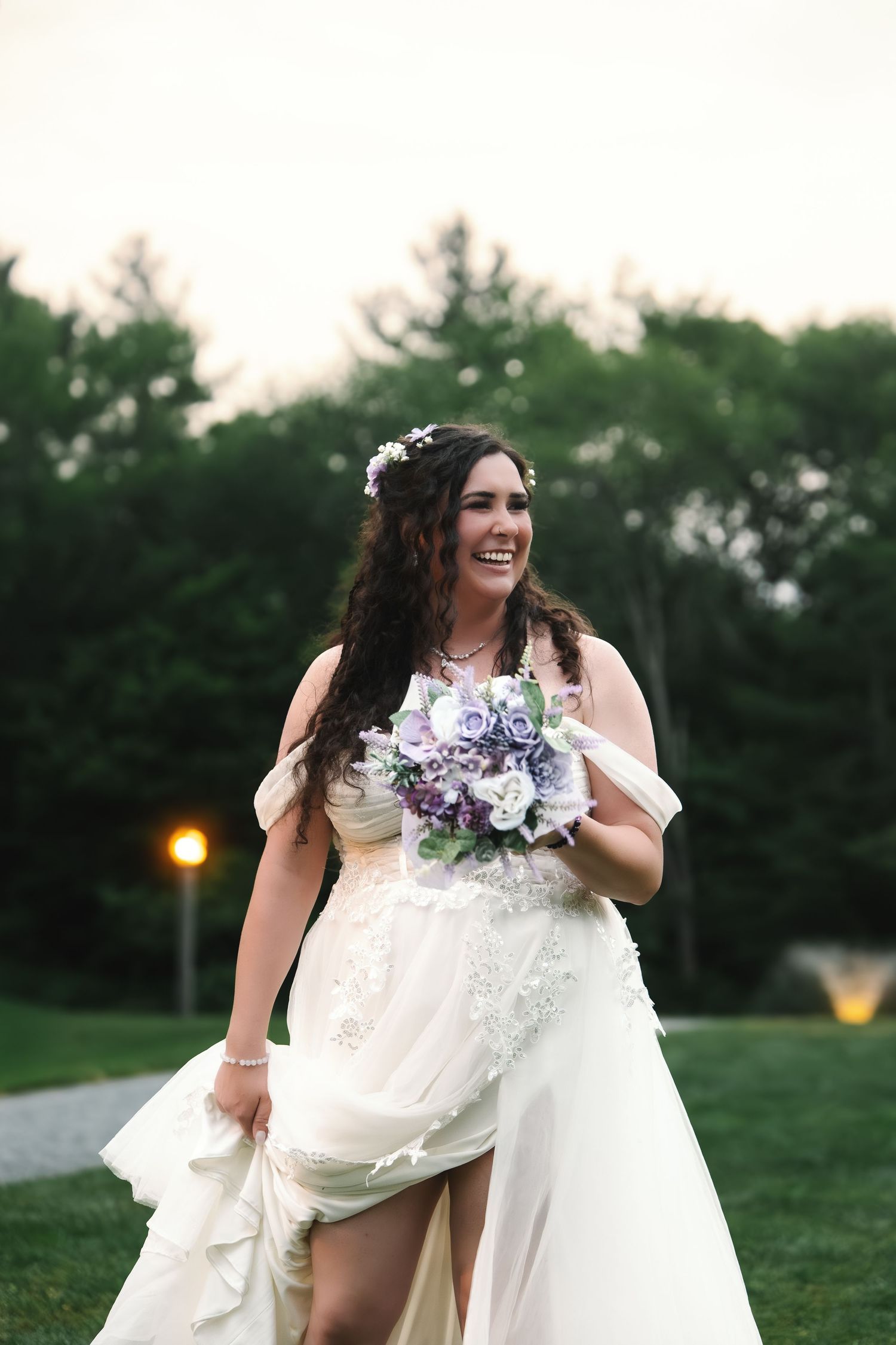 Joyful bride in white lace gown laughing outdoors at dusk holding purple and white bouquet.