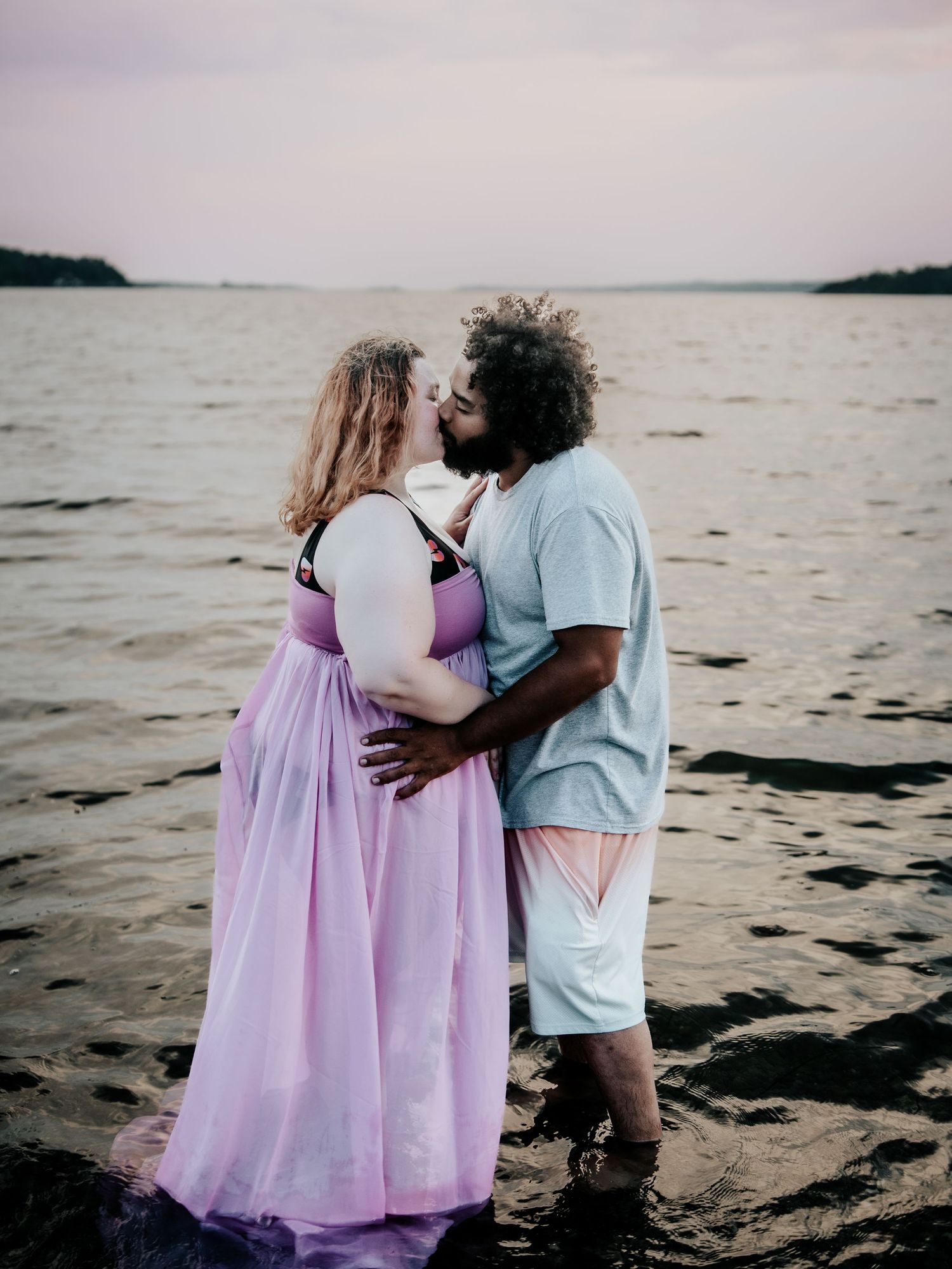 A loving couple kisses while standing in shallow water at dusk, she wears a flowing purple gown.