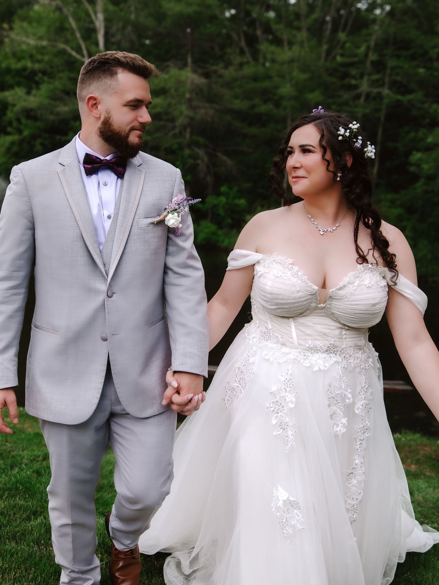 A bride and groom hold hands outdoors, she in a lace off-shoulder gown, he in a gray suit with bow tie.