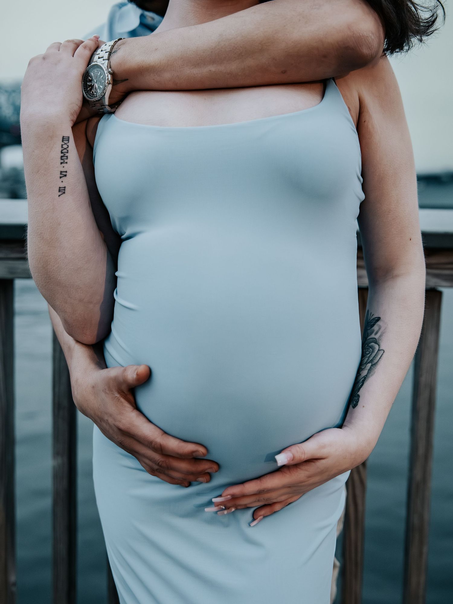 Close-up of a pregnant belly in a light blue dress with a partners tattooed arms lovingly wrapped around her.