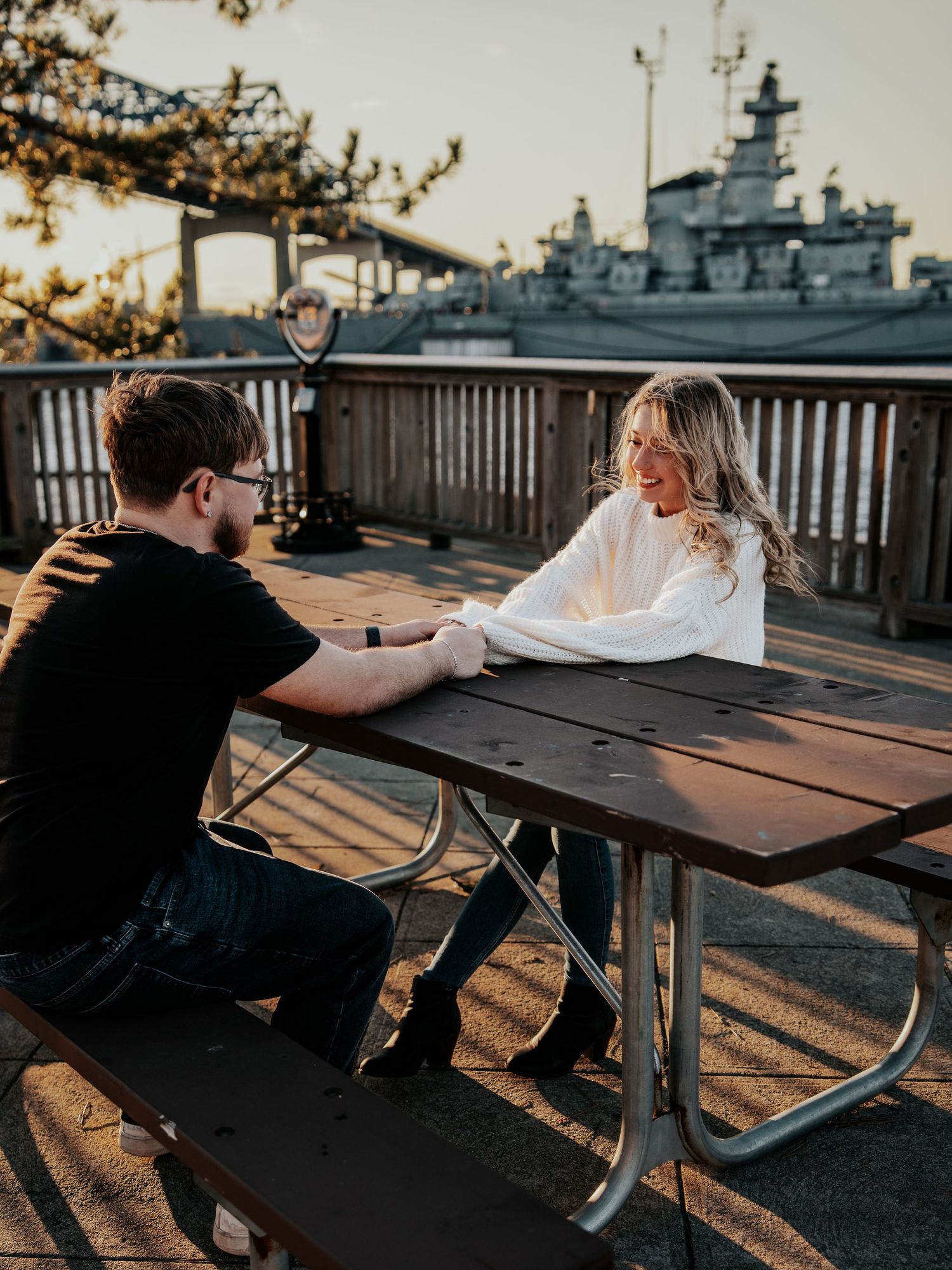 Couple holding hands at an outdoor picnic table near a waterfront with a naval ship in the background.