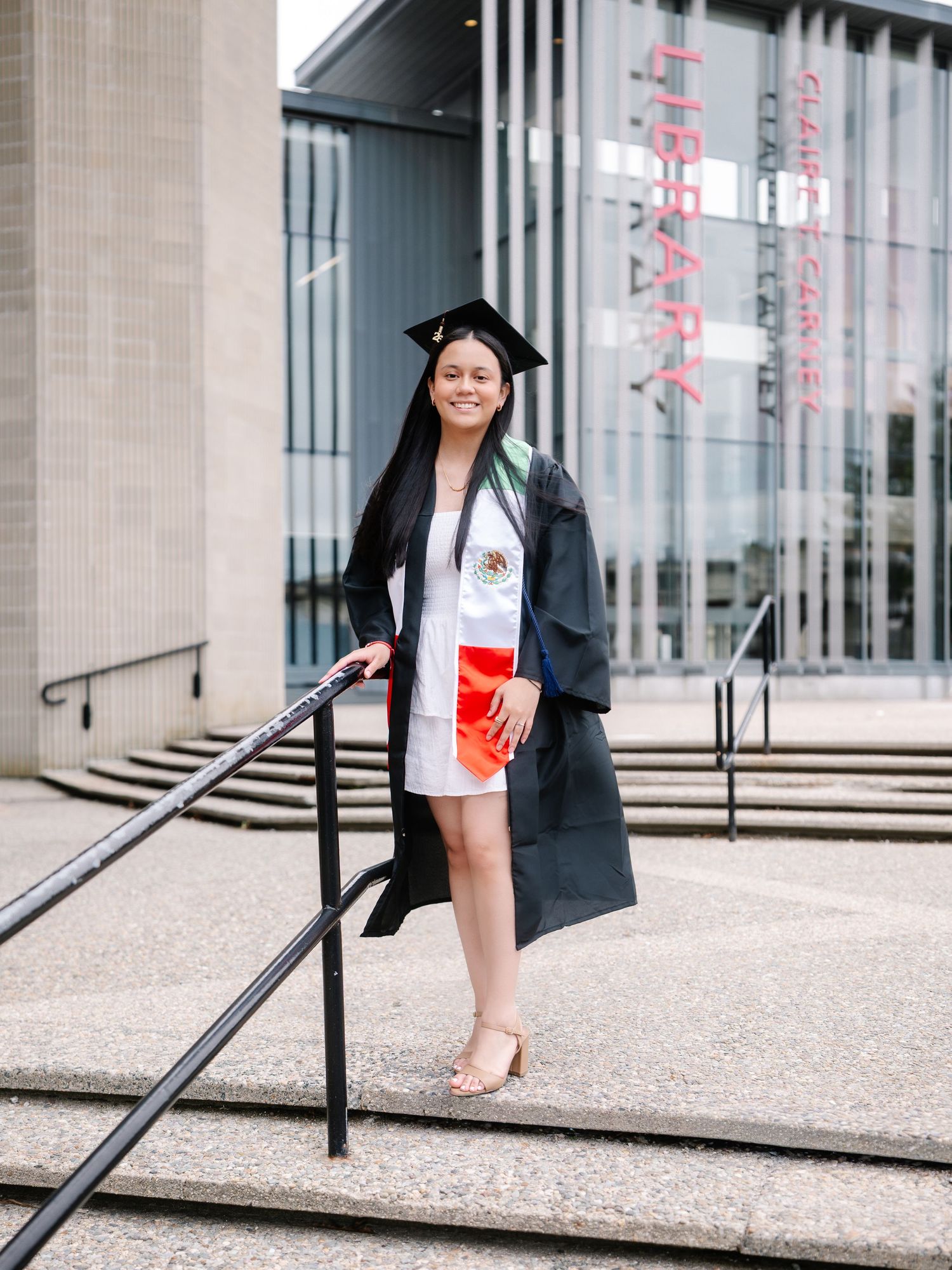 Graduate in black cap and gown with Mexican flag stole posing on steps outside a library.
