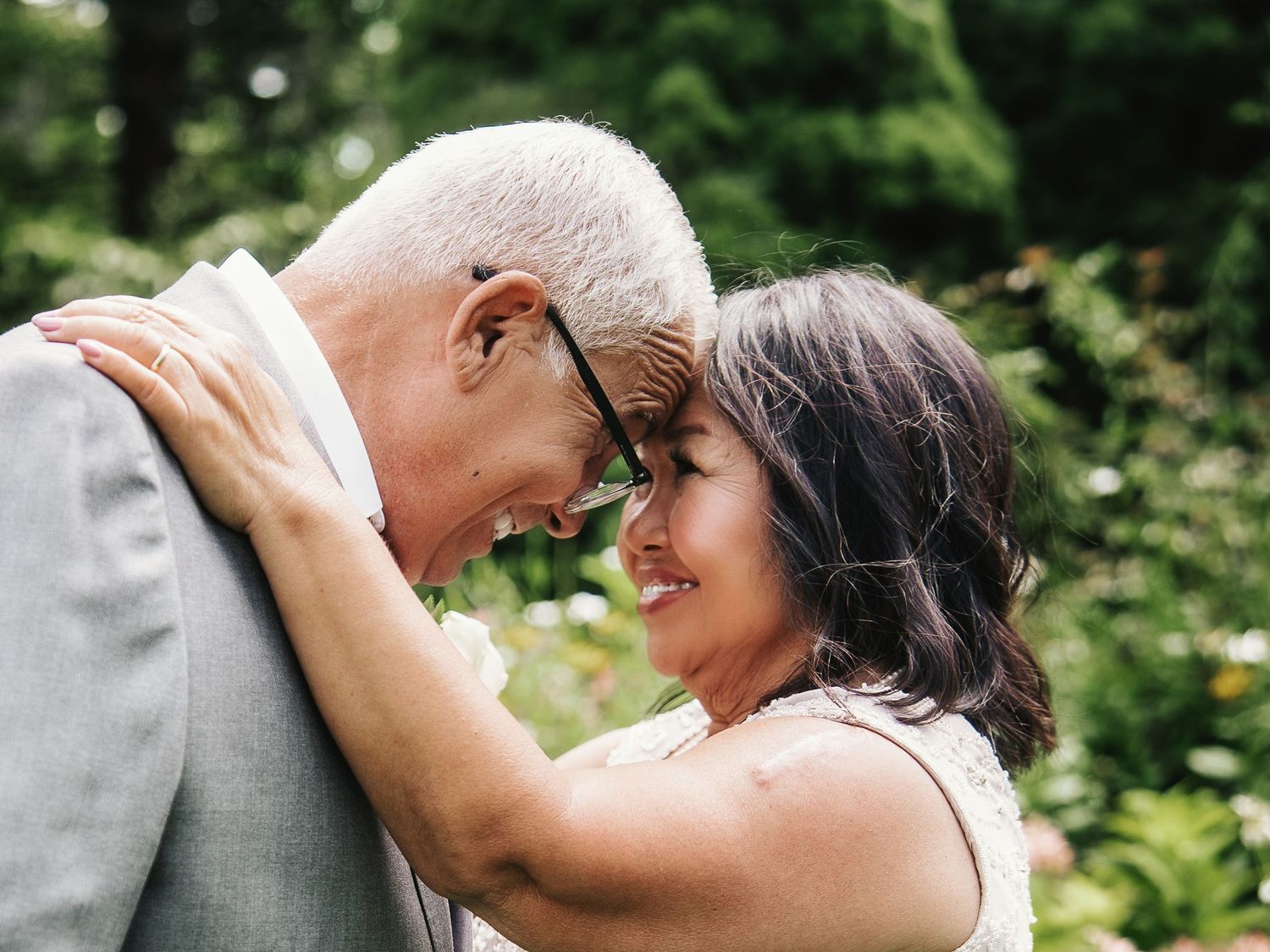 Elderly couple sharing a romantic forehead touch while smiling in a lush green garden setting.