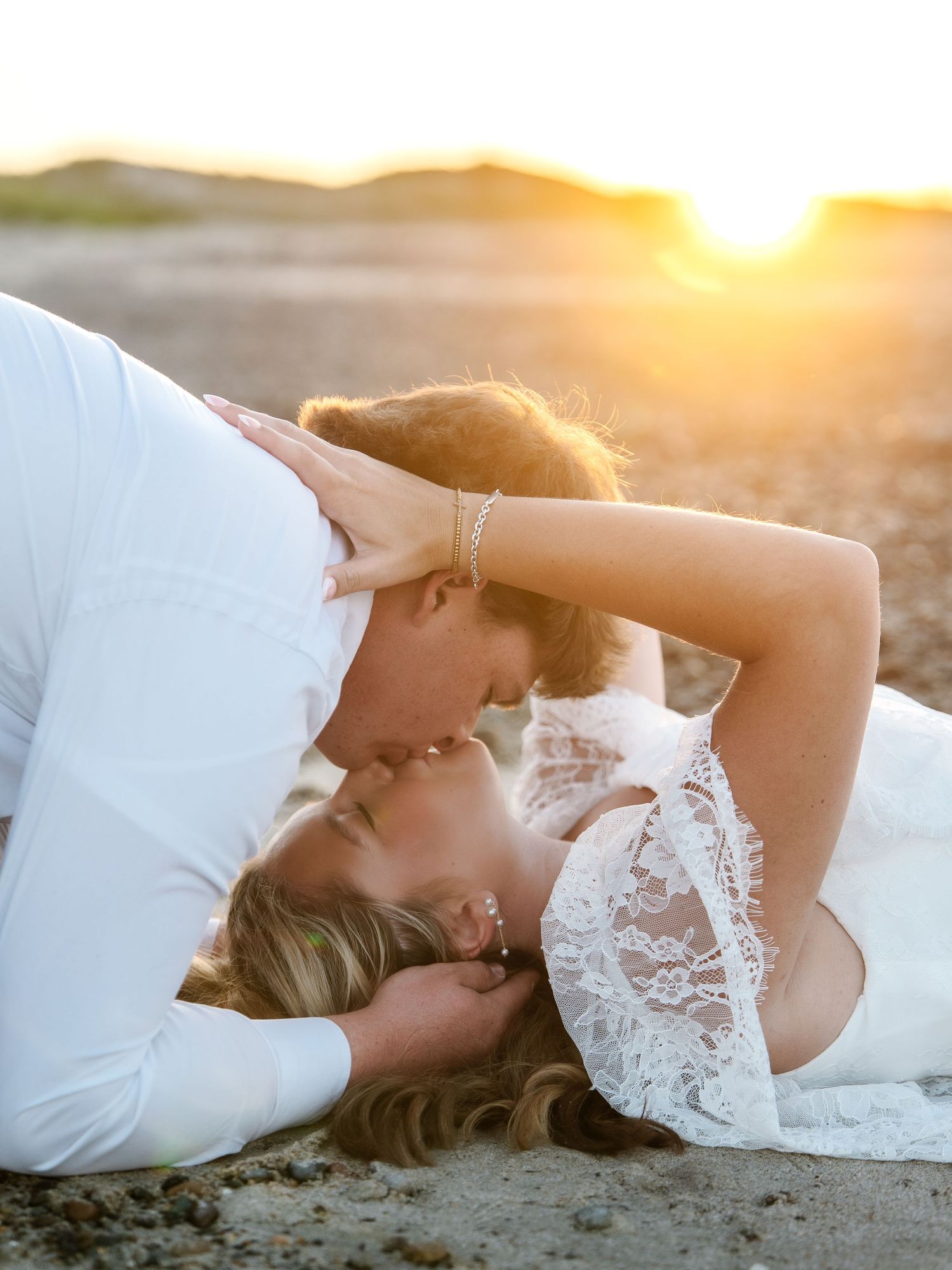 Couple sharing a romantic kiss on the beach at golden sunset, bride in white lace dress lying on sand.