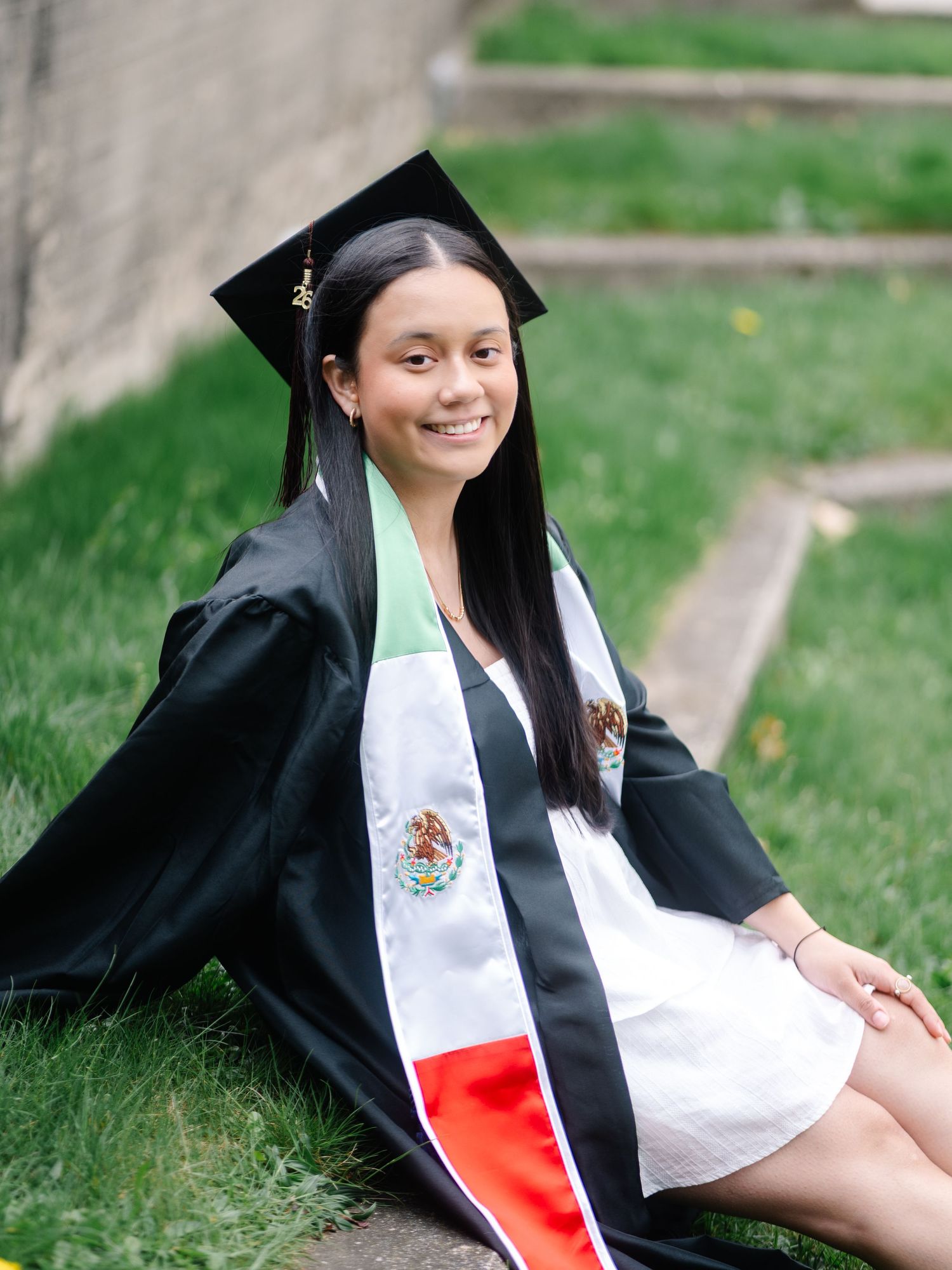 Graduate in black cap and gown with Mexican flag stole sits on grass steps smiling.