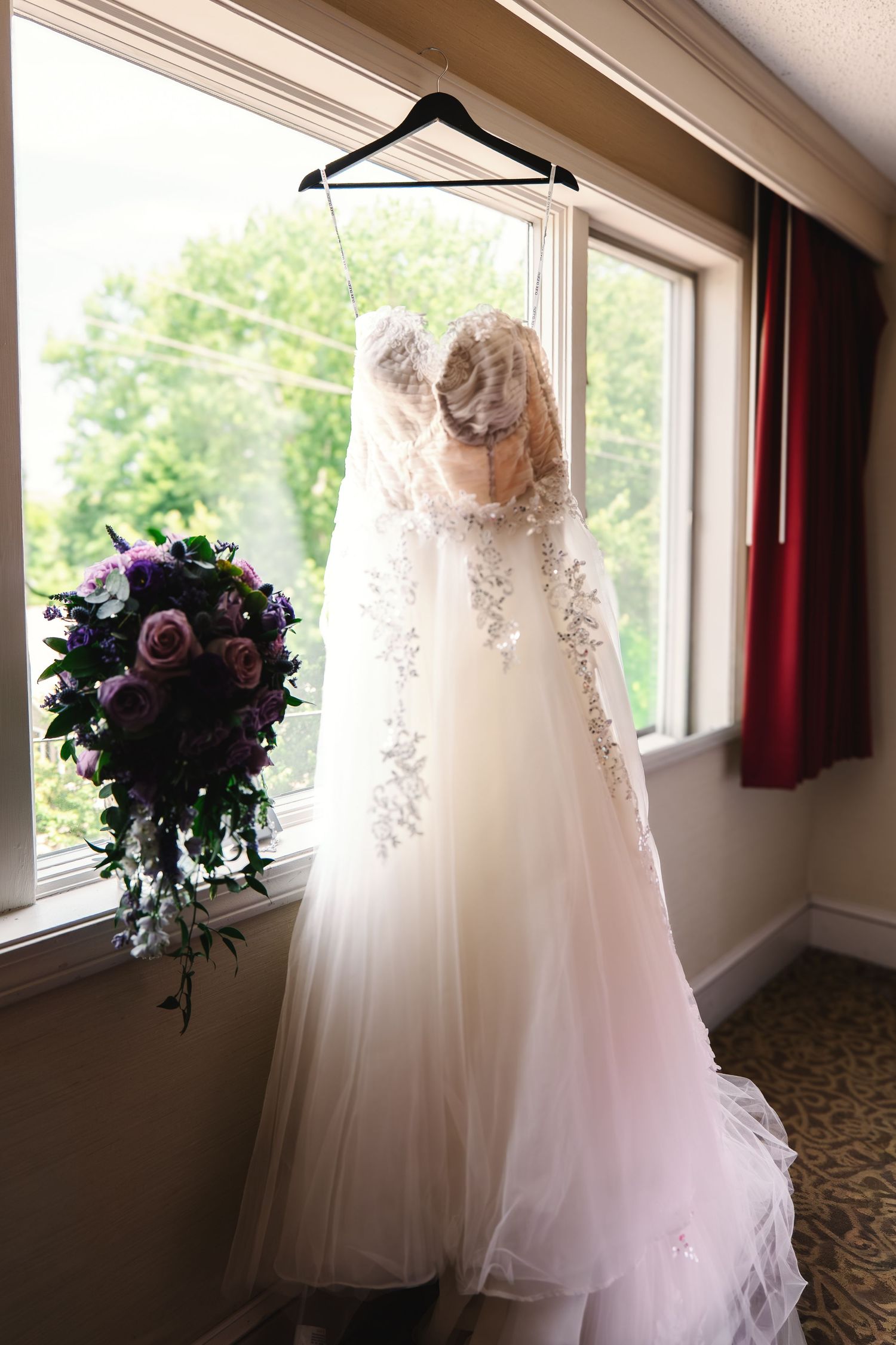 Elegant lace wedding dress hanging by a window with a purple floral bouquet beside it.