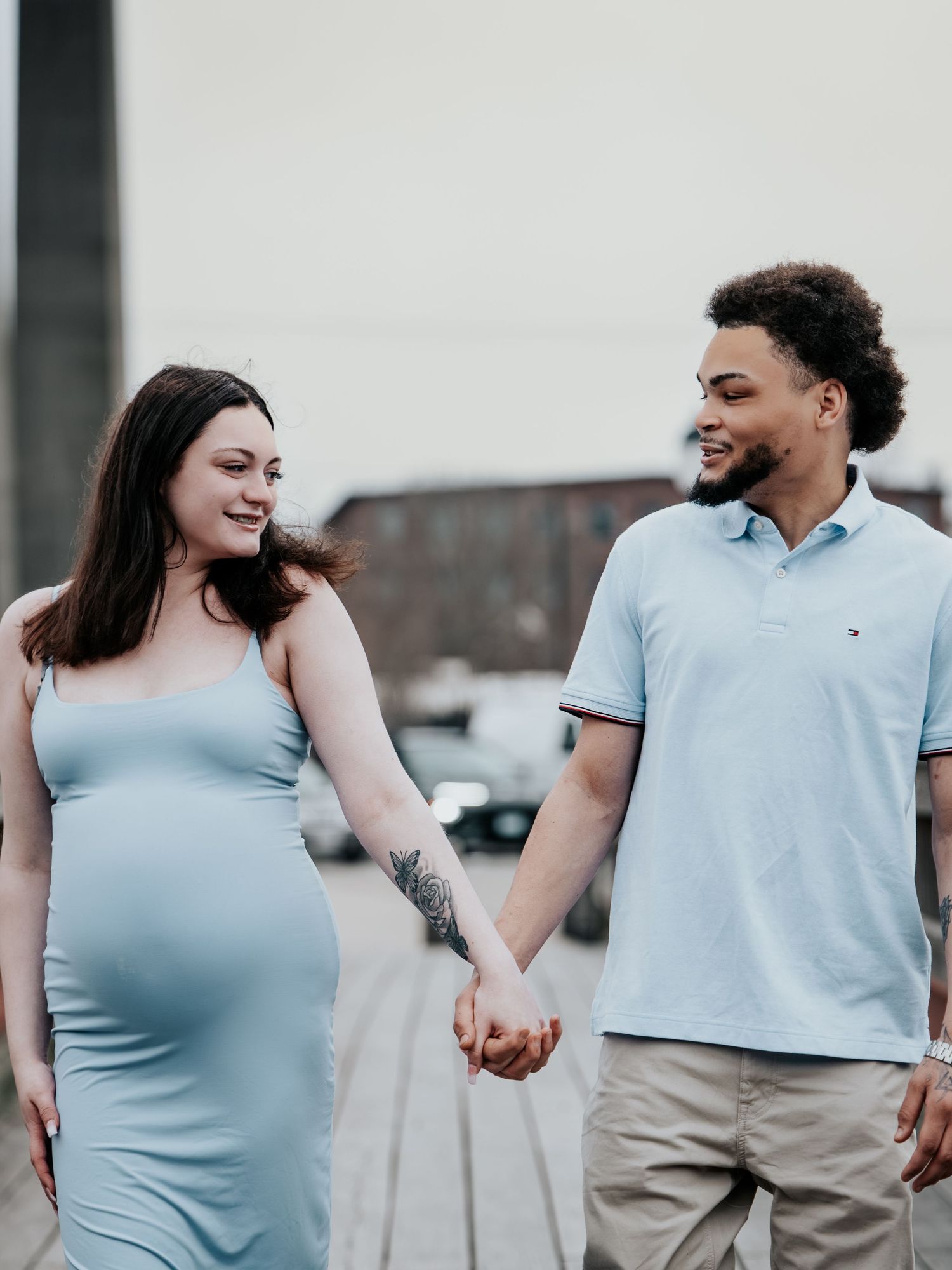 Pregnant woman in light blue dress holds hands with man in polo shirt while walking outdoors together.