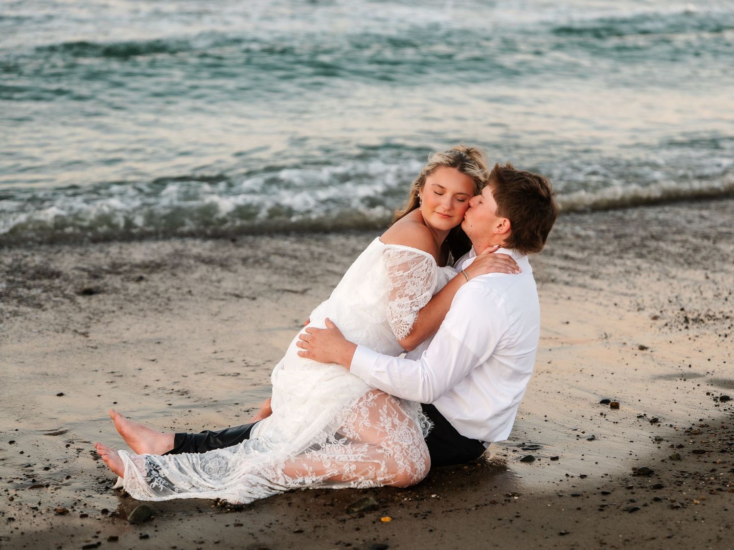 Bride and groom sitting on wet sand at beach, sharing a romantic kiss at dusk.