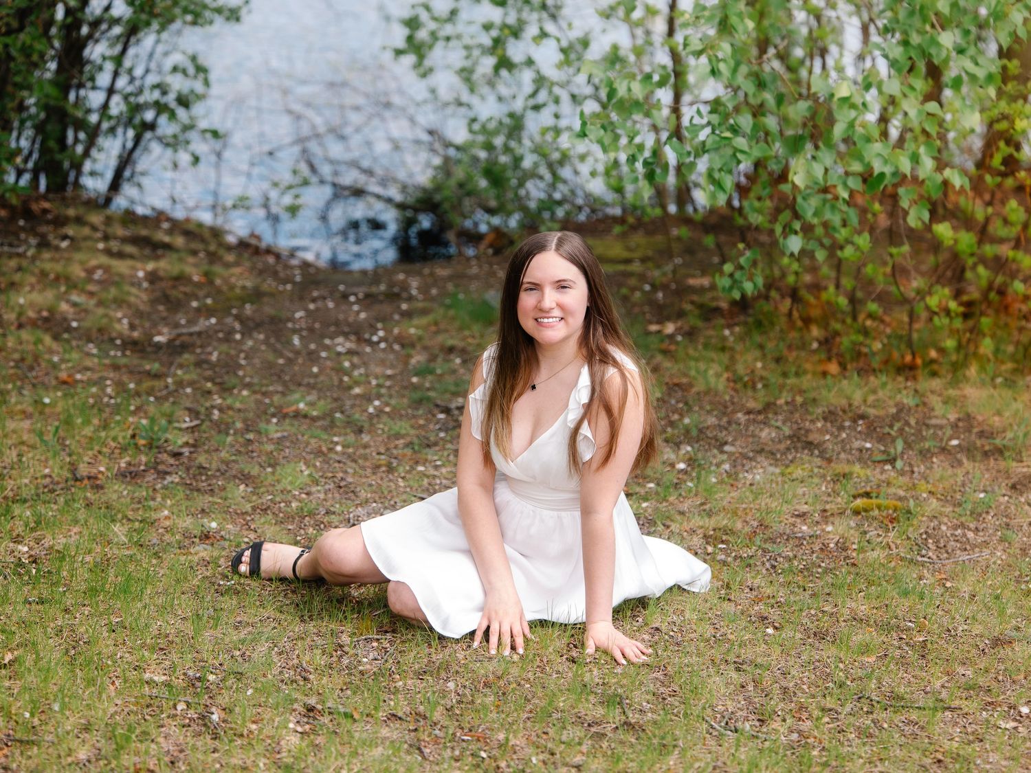 Young woman in white dress sitting on grass near a serene lake surrounded by lush green trees.