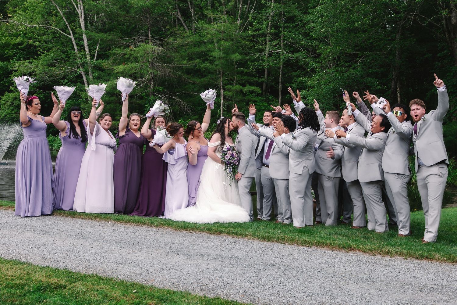 Wedding party celebrating outdoors, bridesmaids in purple gowns and groomsmen in gray suits cheering with bouquets raised.