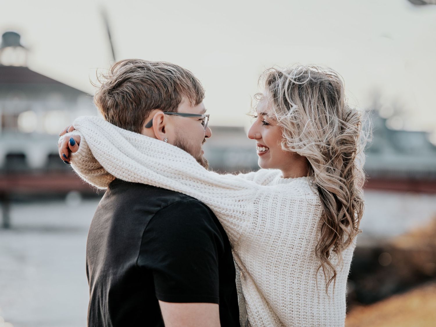 A couple embracing outdoors, smiling at each other, woman in white sweater with arms around man in black shirt.