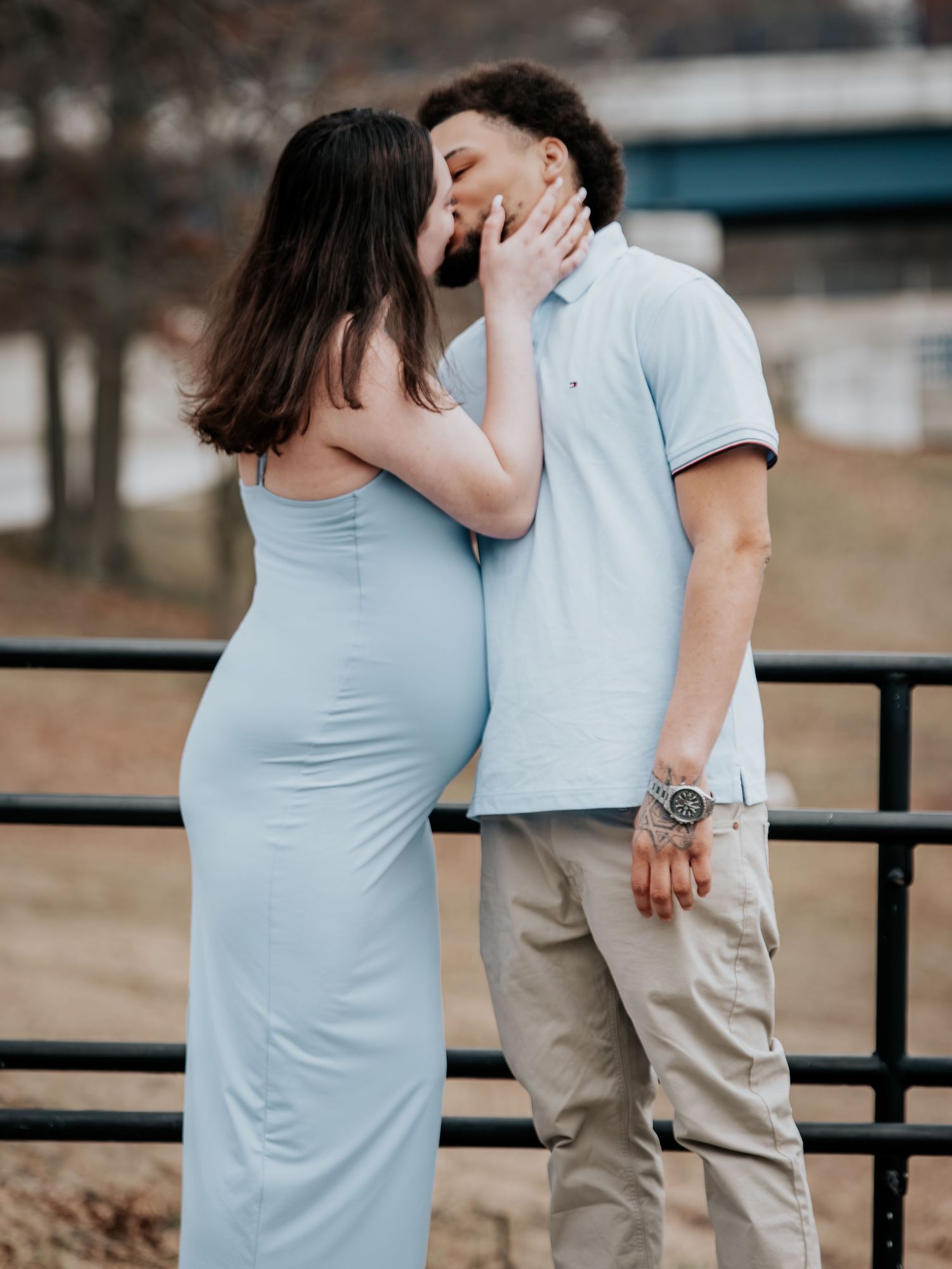 Pregnant woman in light blue dress kissing her partner outdoors near a railing during a maternity photo session.