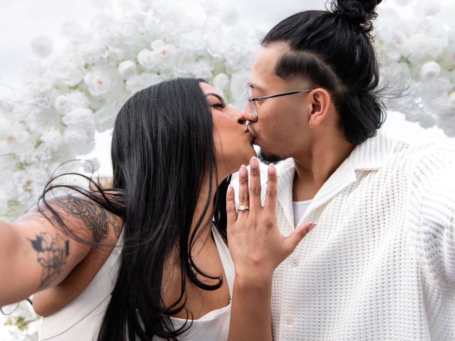 Couple sharing a romantic kiss at a white floral backdrop, woman showing engagement ring, wearing white outfits.