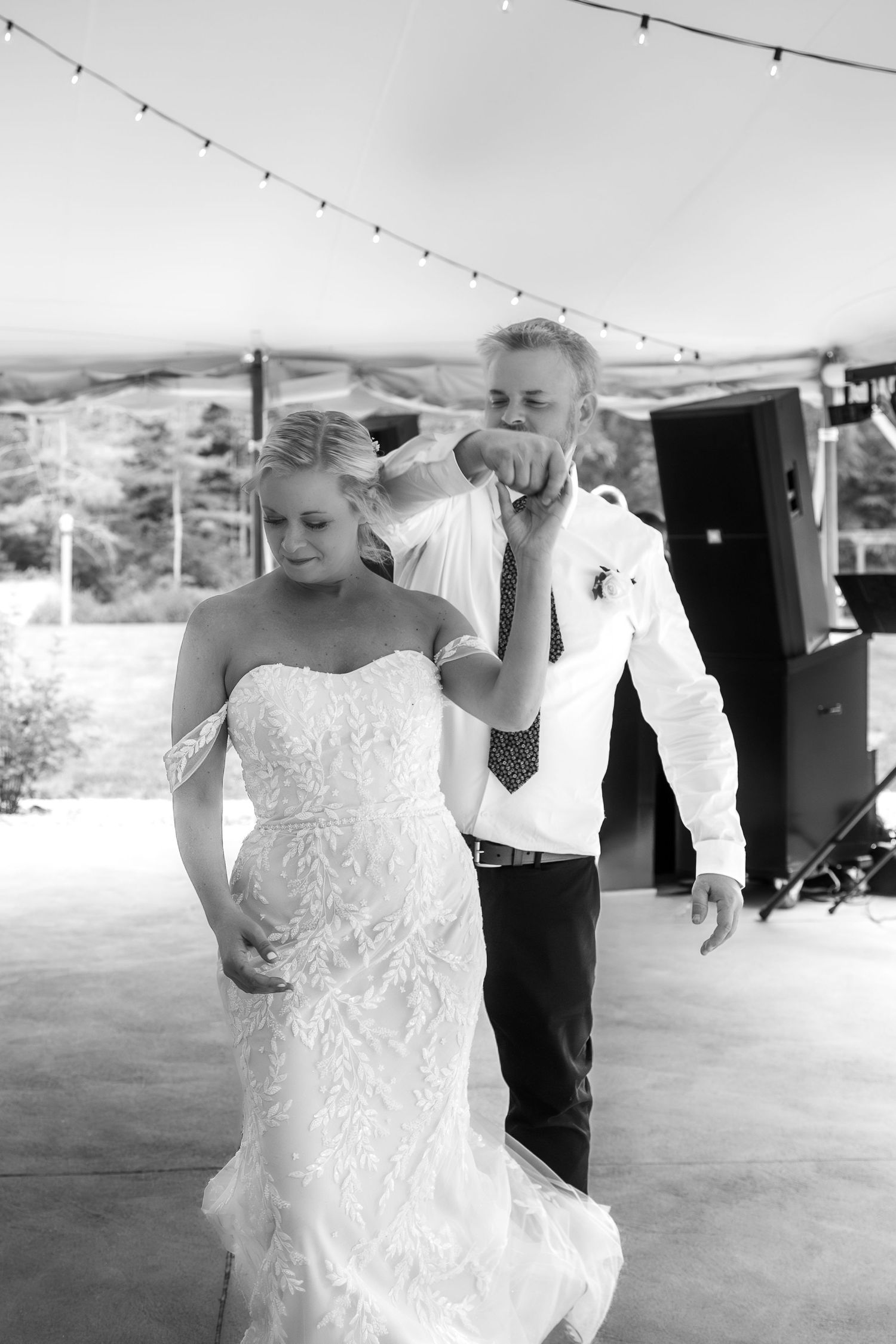 Black and white photo of bride and groom sharing first dance at outdoor wedding reception under string lights.