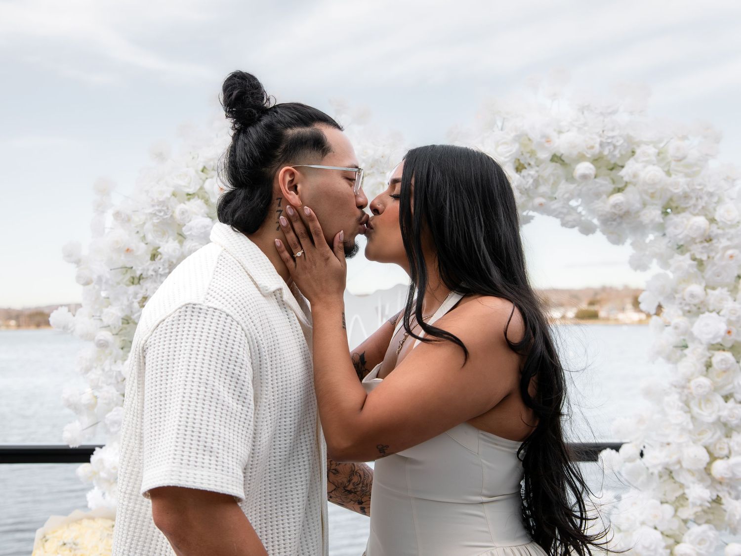 Couple sharing a romantic kiss in front of a white floral arch by the water during an engagement or wedding.