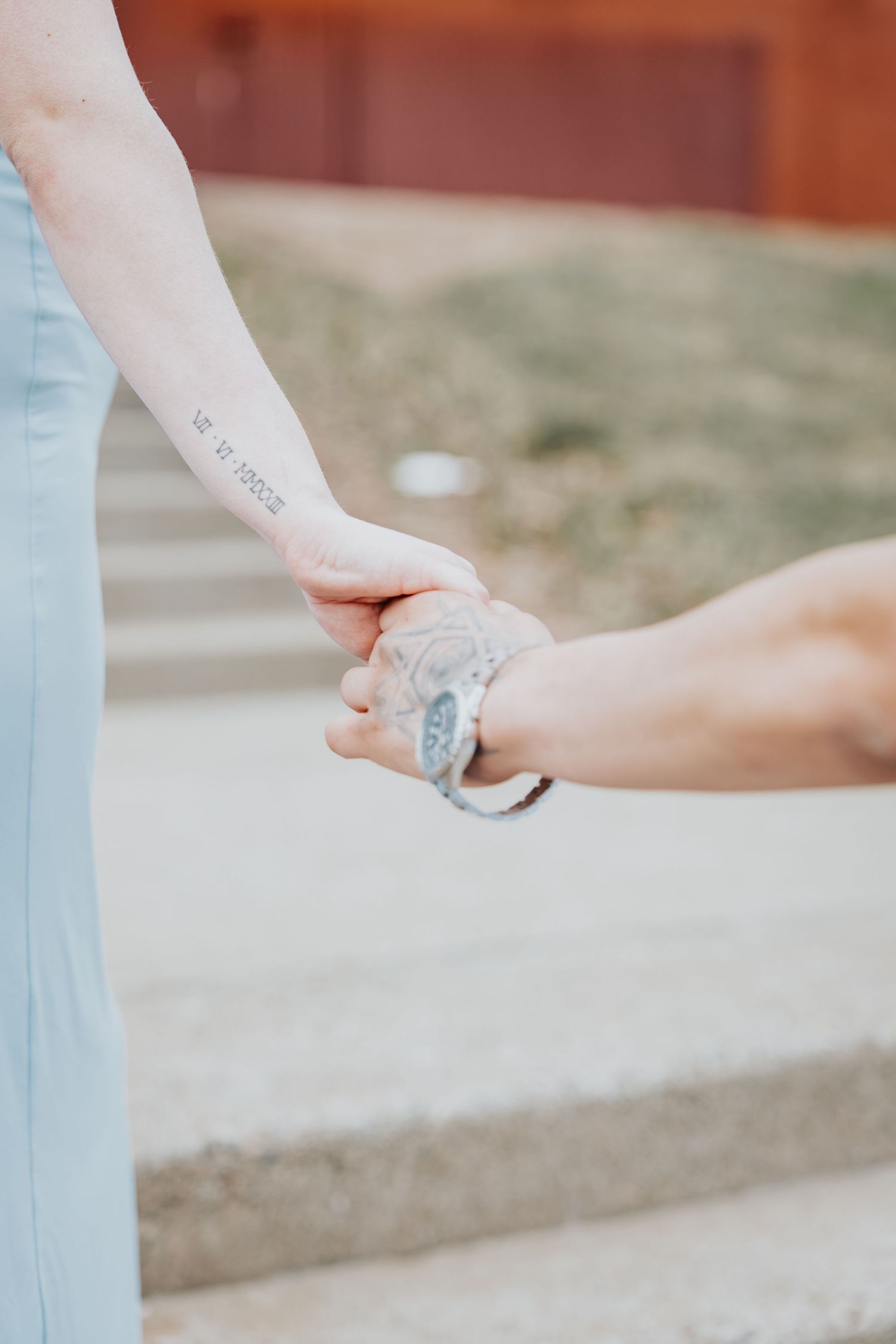 Two people holding hands outdoors, one with a forearm tattoo and the other with a tattooed wrist and bracelet.