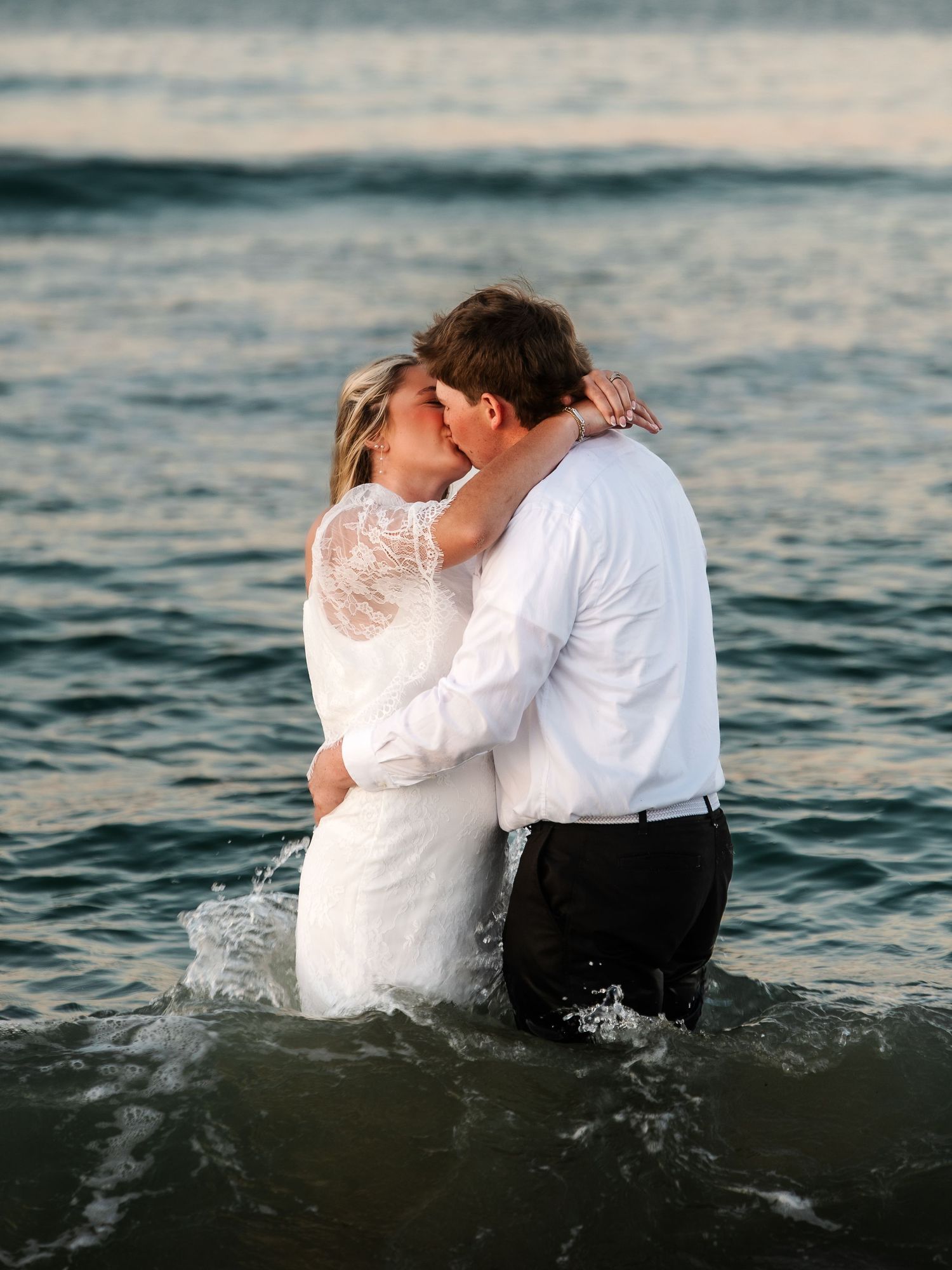 Newlyweds sharing passionate kiss while standing waist-deep in ocean waves during beach wedding shoot.