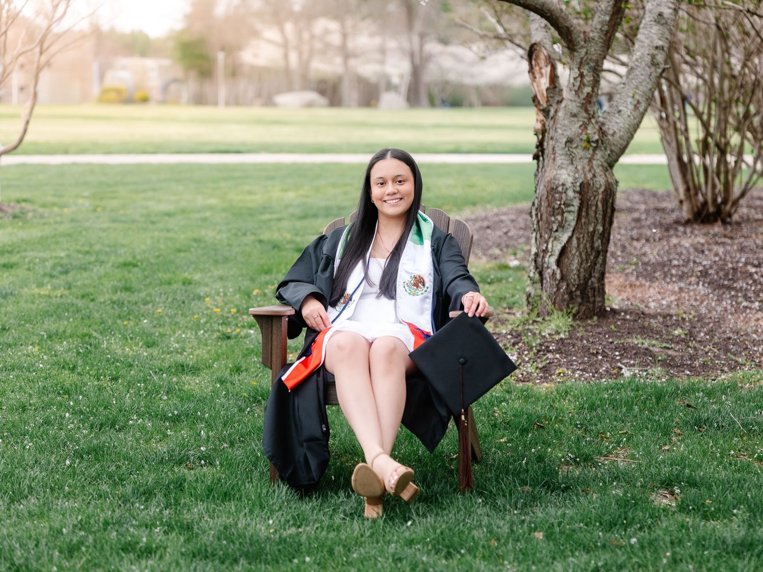 A smiling graduate in black cap and gown sits on a wooden bench outdoors, holding a diploma and graduation cap.