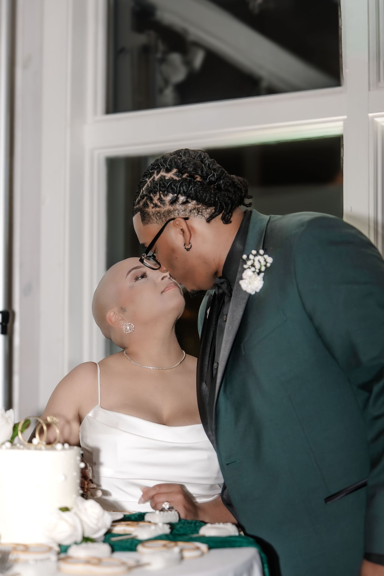 Newlywed couple sharing a kiss at their wedding cake cutting ceremony, groom in green suit with boutonniere.
