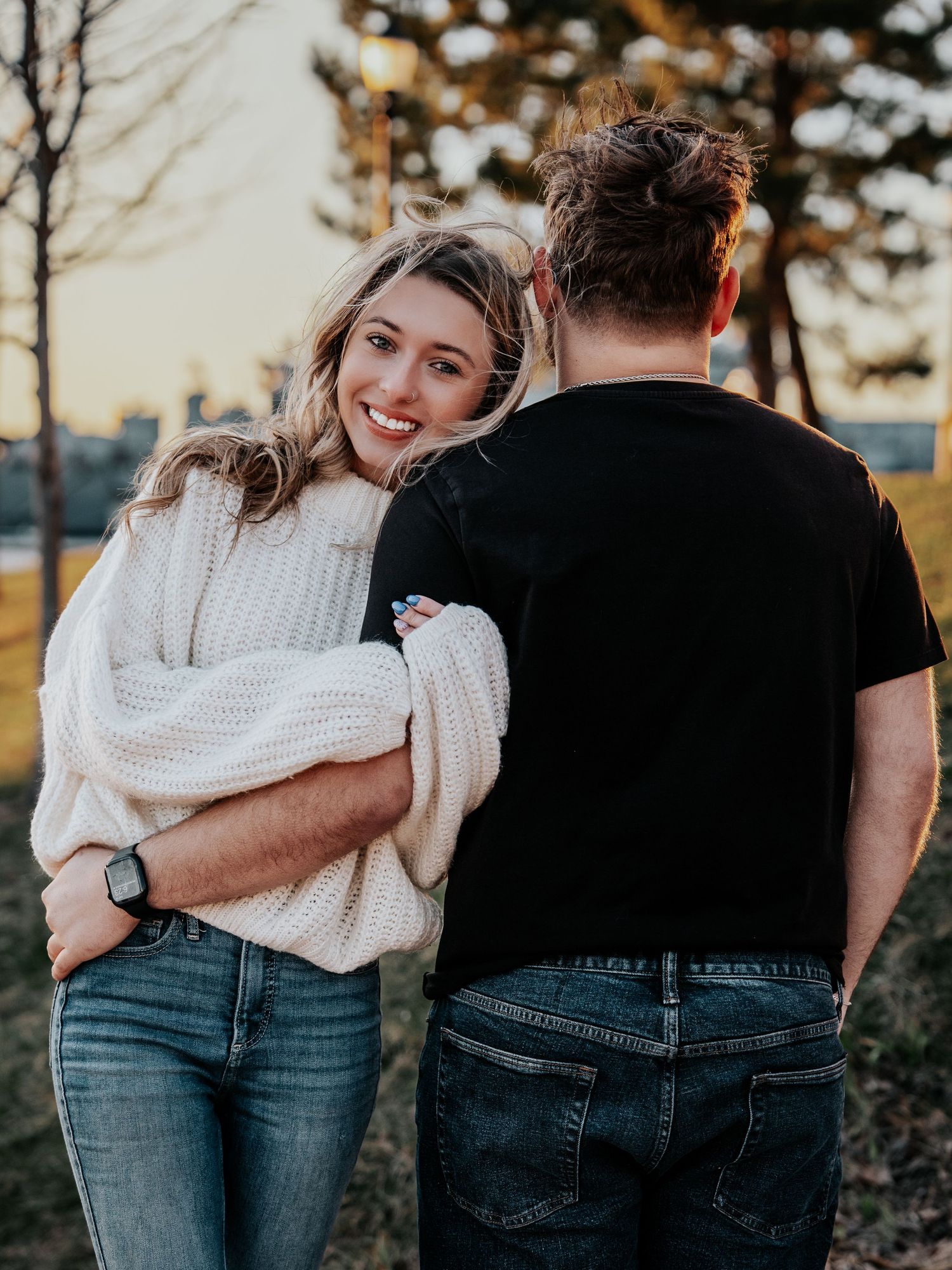 Smiling blonde woman in white sweater hugs man in black t-shirt from behind at sunset park.