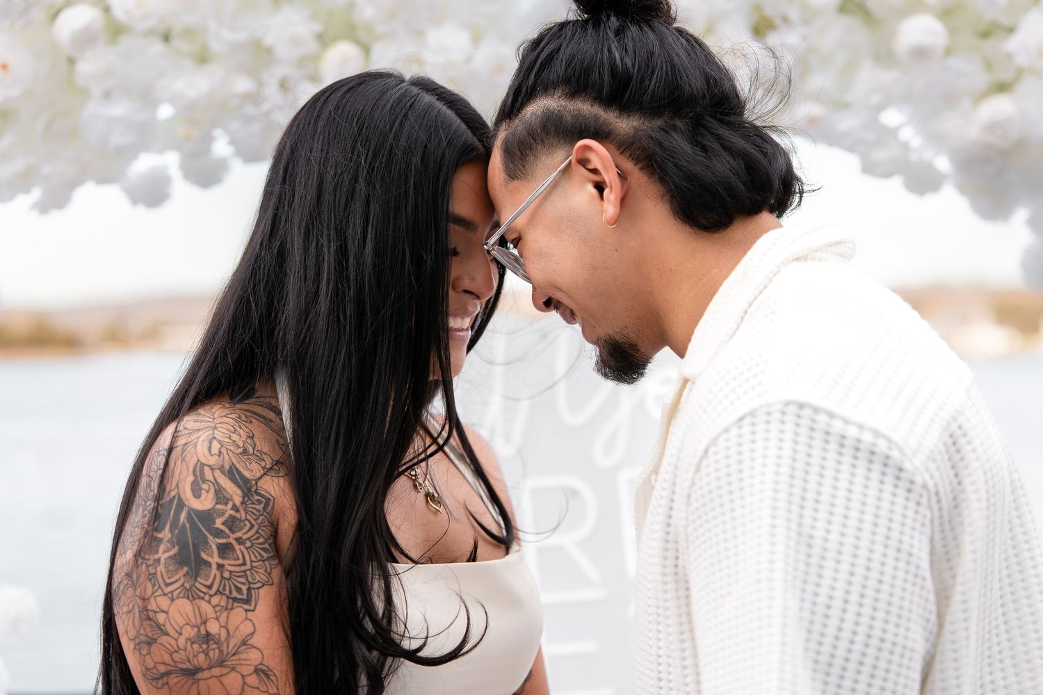 Couple with tattoos sharing an intimate moment, touching foreheads in a romantic outdoor setting with white floral decor.