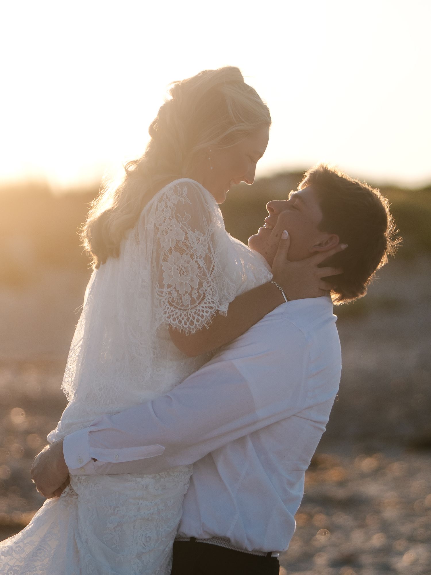 Groom holding bride in white lace dress as they share a romantic moment during beach sunset.