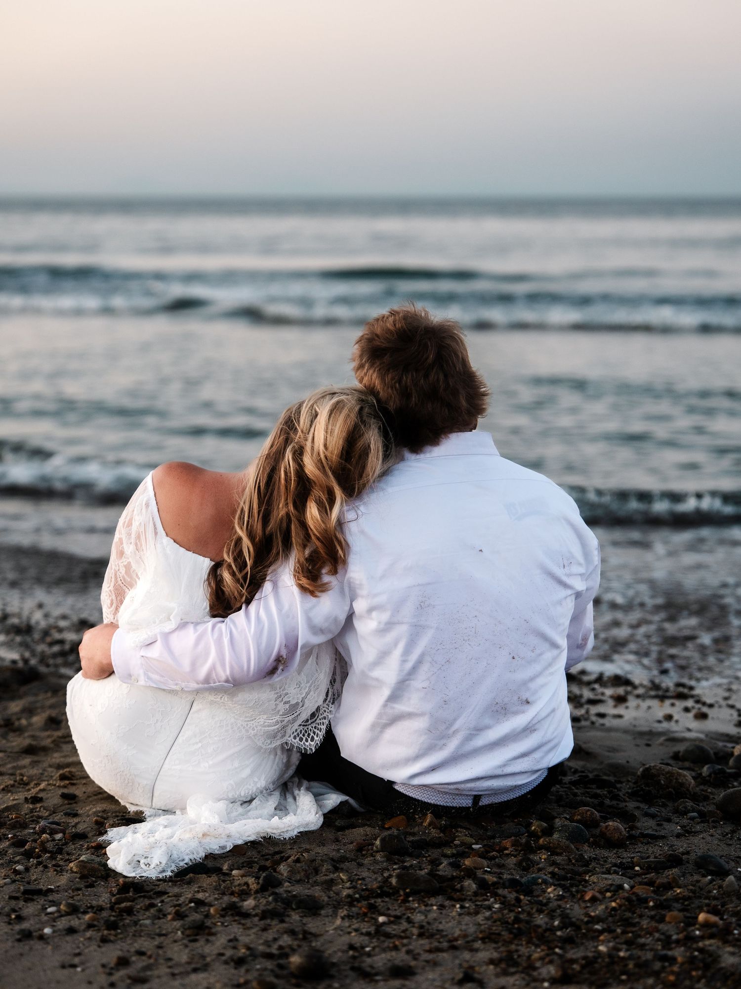 Bride and groom sitting together on beach sand, embracing while gazing at the ocean.
