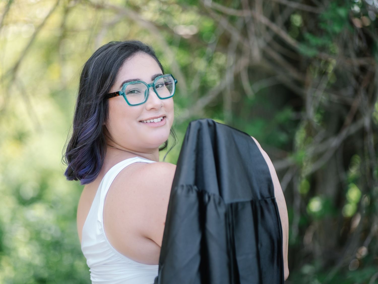 Smiling young woman in white dress carrying black graduation gown over shoulder outdoors in nature.
