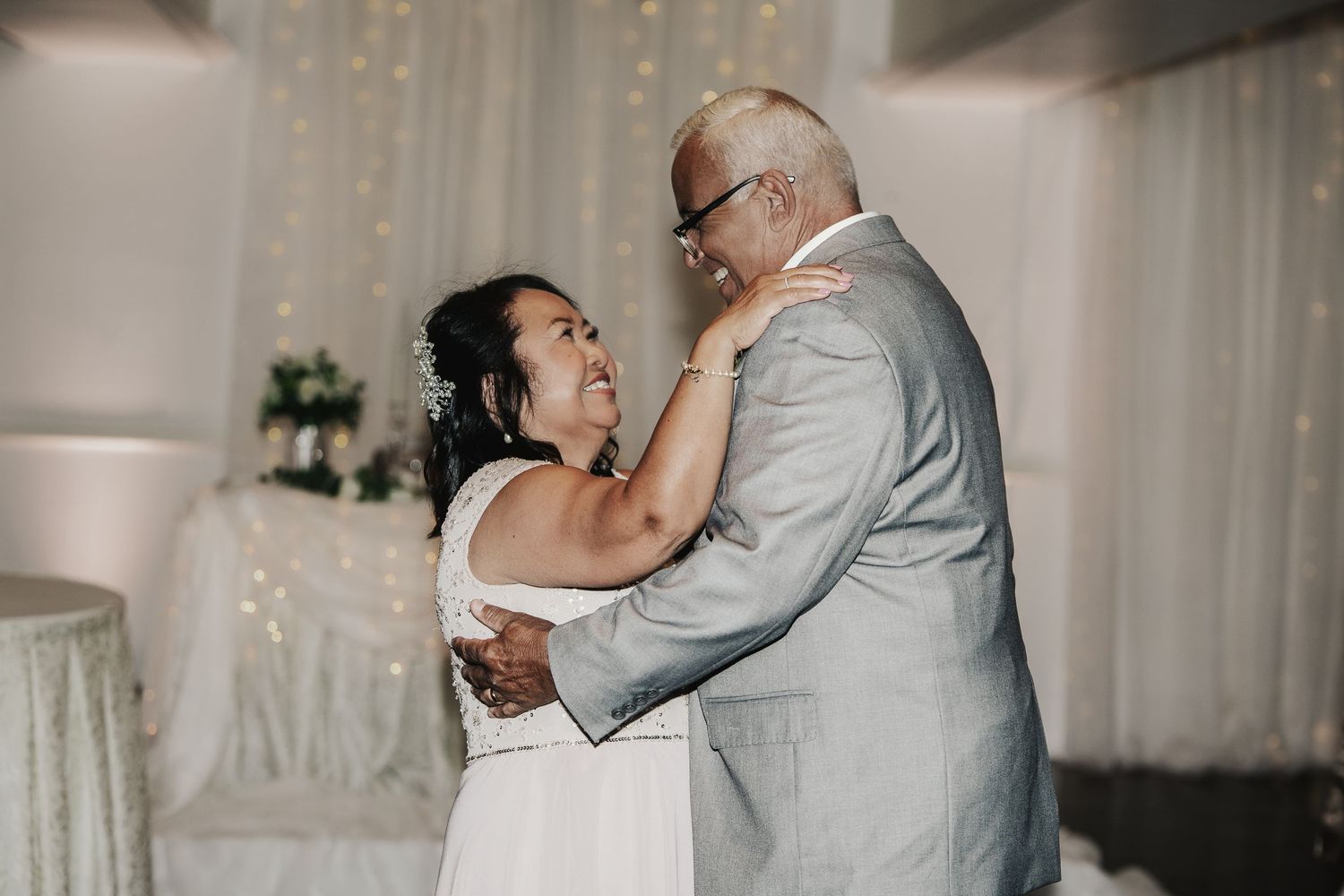 Couple shares first dance at wedding reception with fairy light backdrop, bride in white gown embracing groom in gray suit.