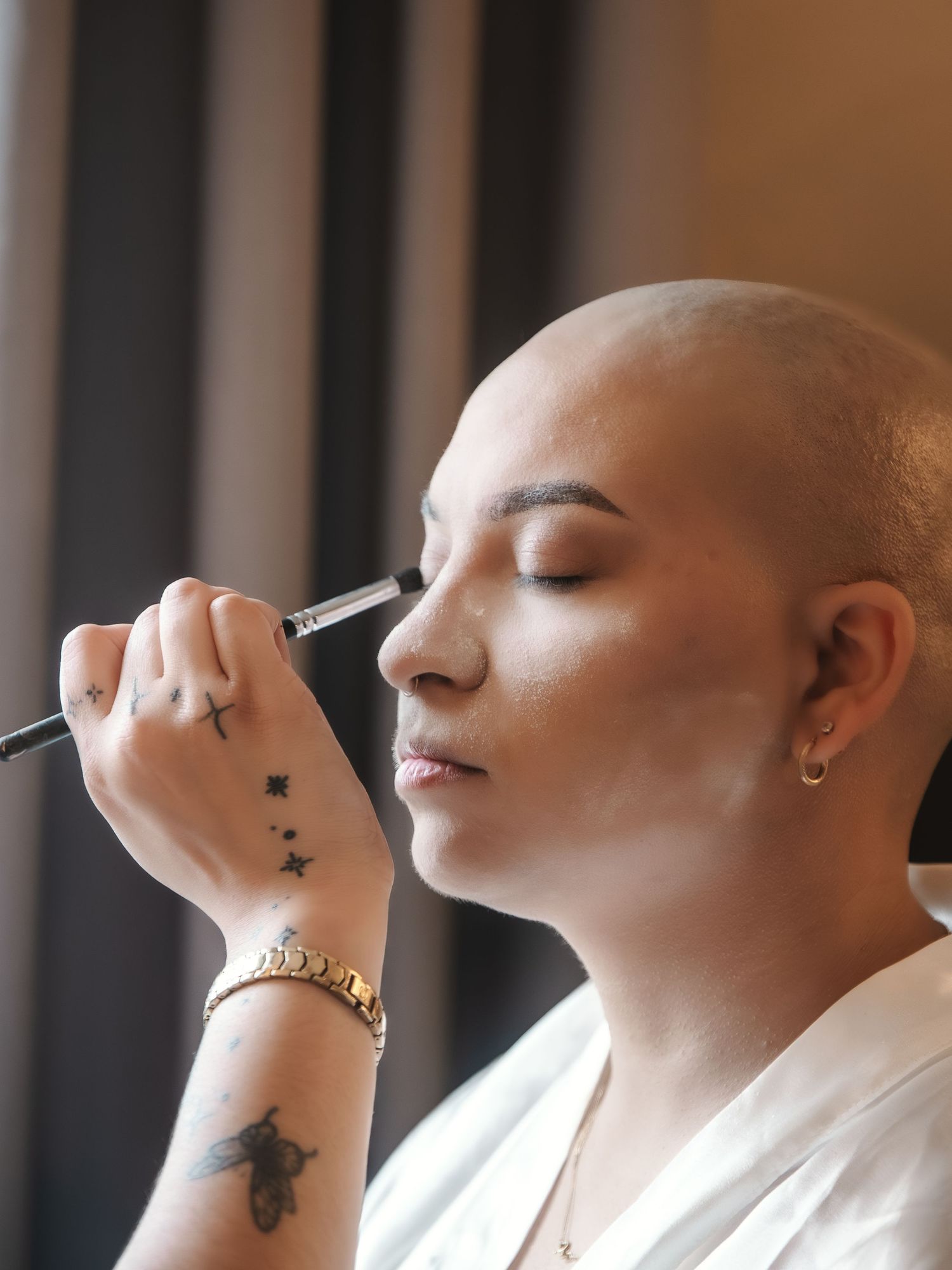 A bald woman with tattoos gets eye makeup applied with a brush, wearing a gold bracelet and white top.